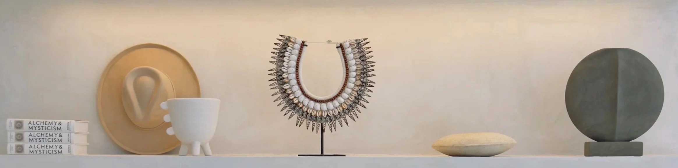 Decorative shelf with books titled 'Alchemy & Mysticism', a beige circular wall plaque with a white ear-shaped object, a white ceramic vase with three legs, a tiered necklace with shells and pointed metal accents, a small beige dish, and a large gray spherical sculpture on a pedestal.