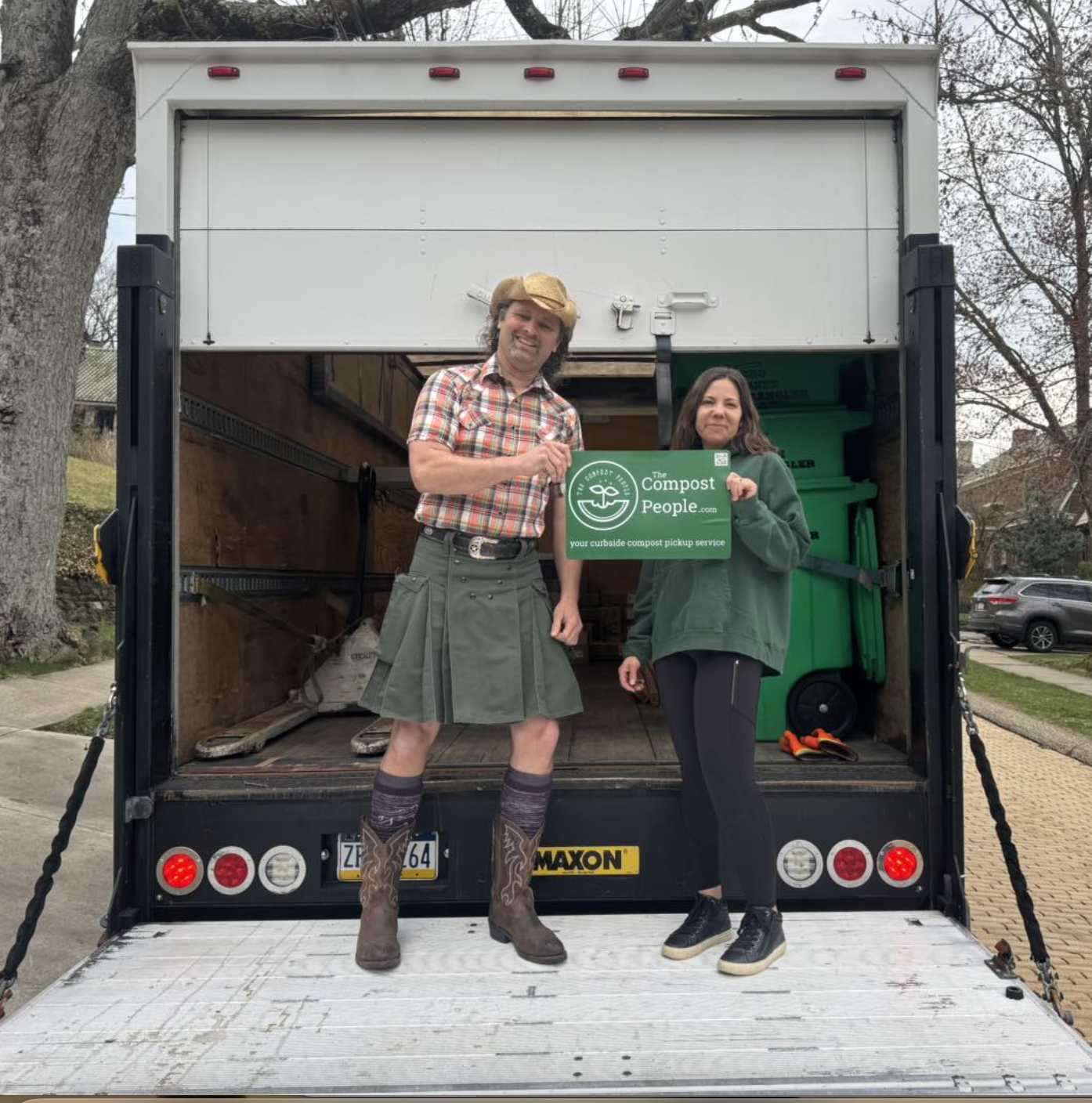 A man and woman standing inside a moving truck, holding a green sign that reads 'The Compost People: your curbside compost pickup service'. The man is dressed in a plaid shirt, kilt, and cowboy hat, with knee-high socks and boots. The woman is dressed casually in a green hoodie and black pants. There are green recycling bins and other equipment inside the truck.