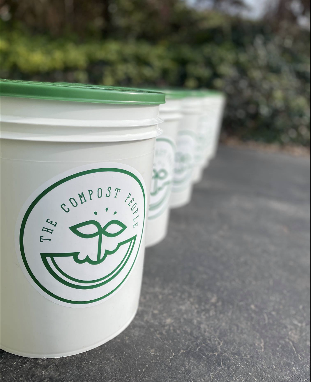 Three white buckets with green lids and logo, placed on a paved surface outdoors with greenery in the background.