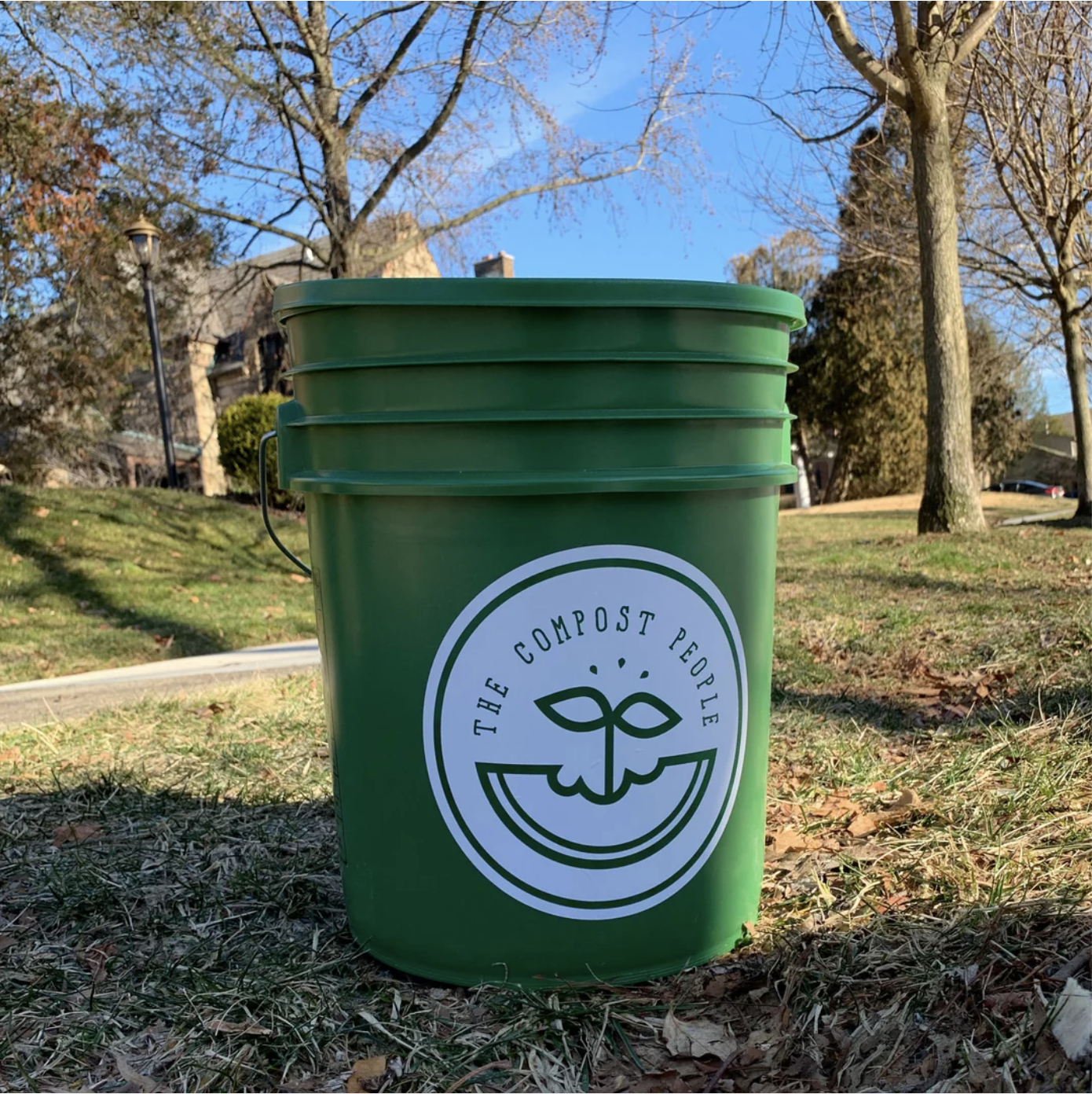 Green compost bin labeled 'The Compost People' with a smiling face logo, outdoors on a grassy area with trees and houses in the background during daytime.