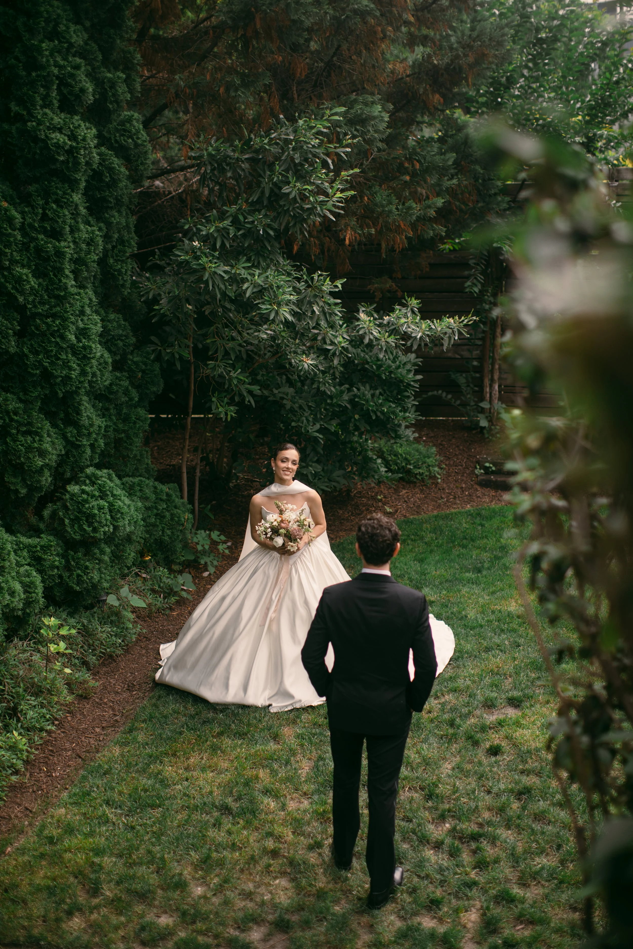 A bride in a wedding gown holding a bouquet of flowers and a groom dressed in a black suit walking towards her in a lush garden with tall trees and shrubs.
