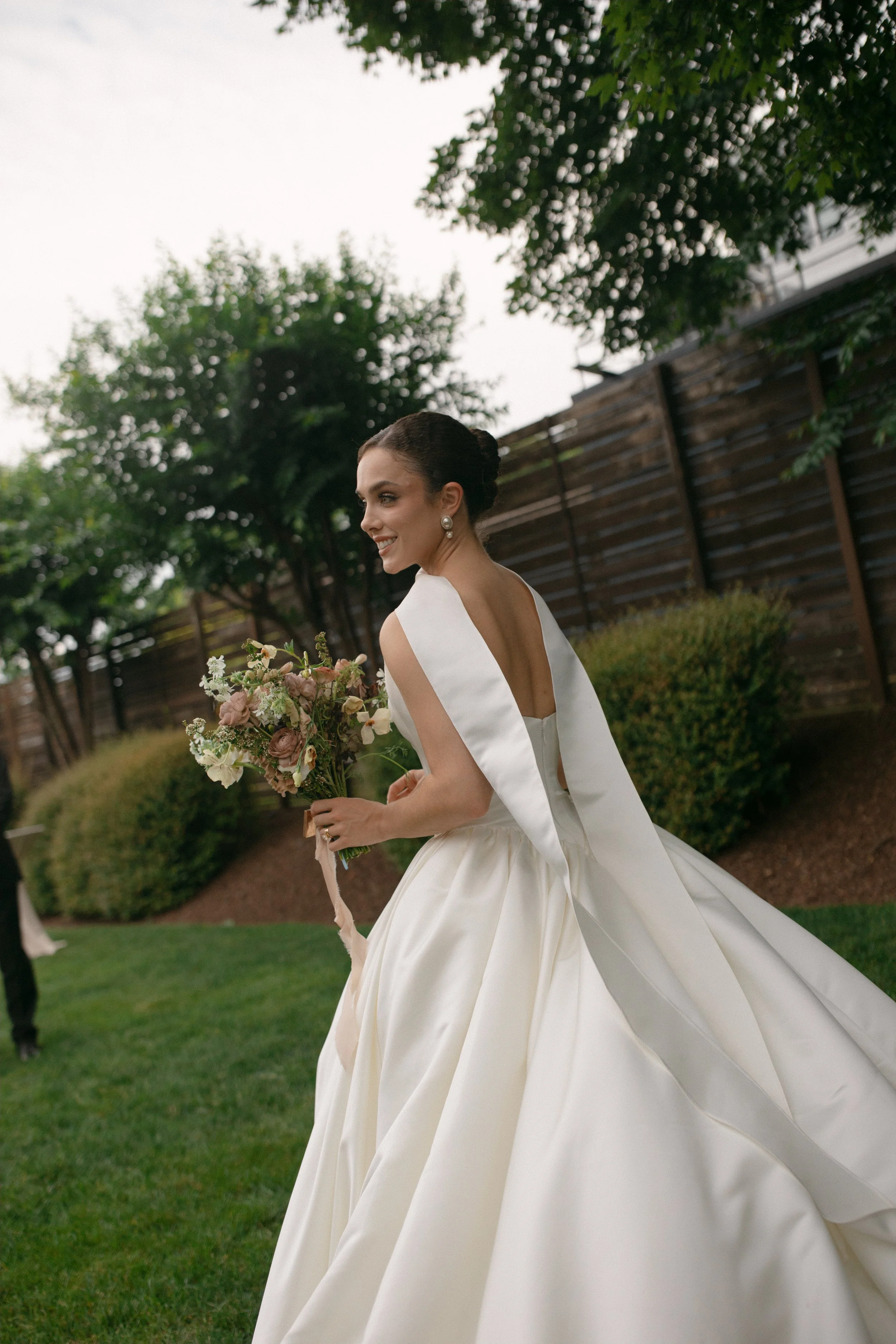 A bride in a white wedding gown with a satin finish, holding a bouquet of flowers, smiling in an outdoor garden setting with trees and a wooden fence.