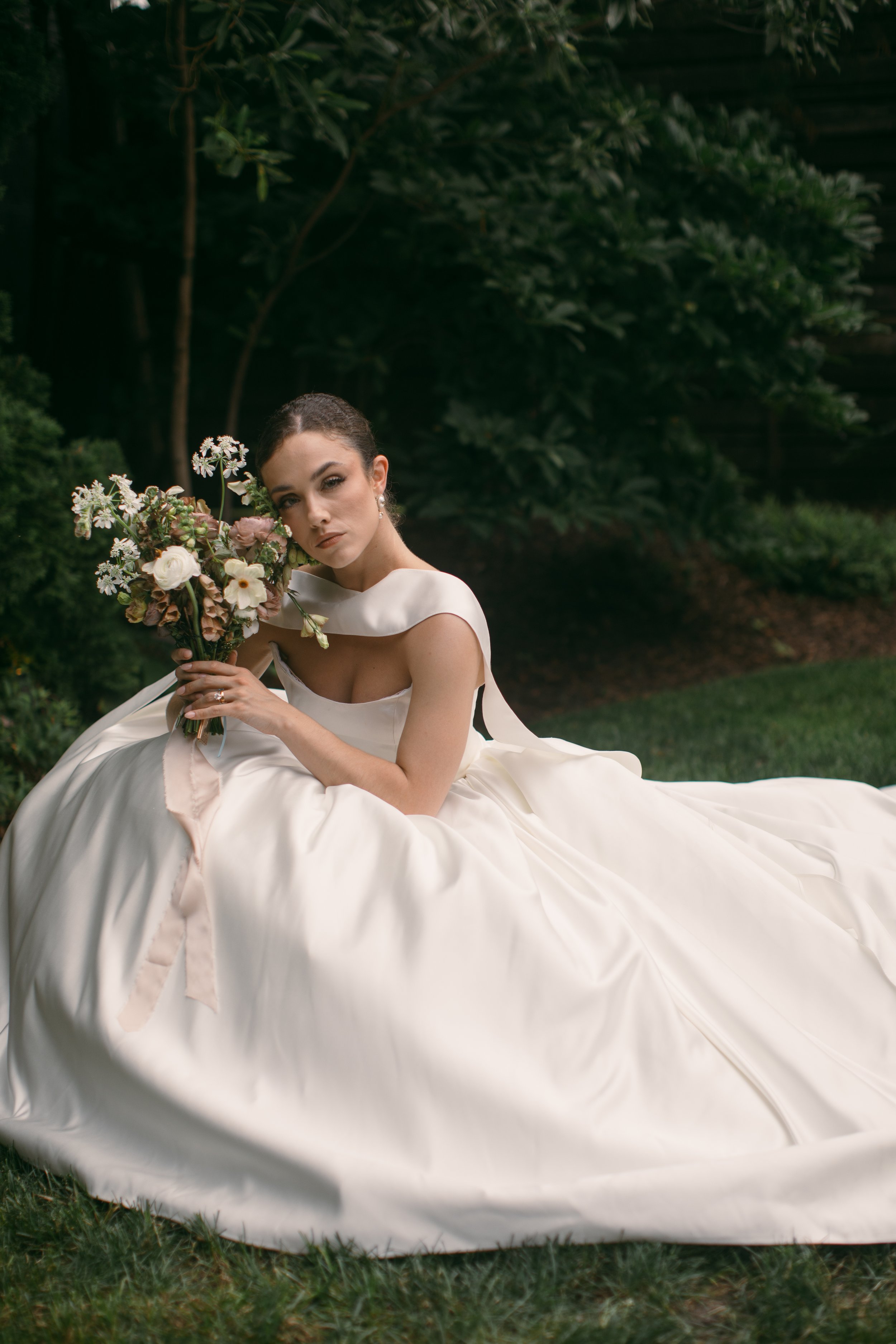 A woman in a white wedding dress is sitting outdoors on grass with trees and bushes in the background, holding a bouquet of flowers, and looking at the camera.