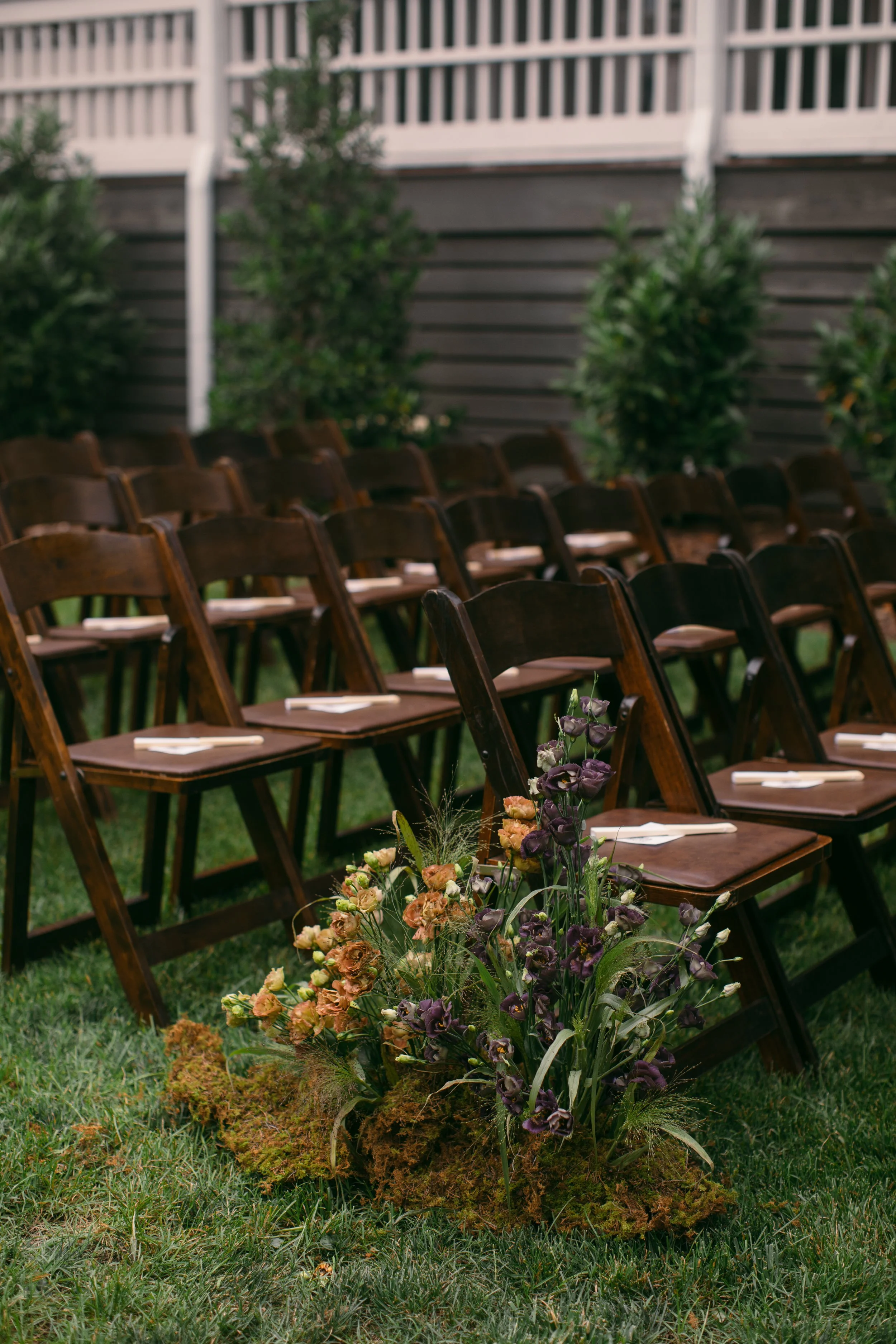 A row of empty wooden chairs set outdoors with white napkins on each, facing a garden with greenery and a gray fence, and a floral arrangement in the foreground.