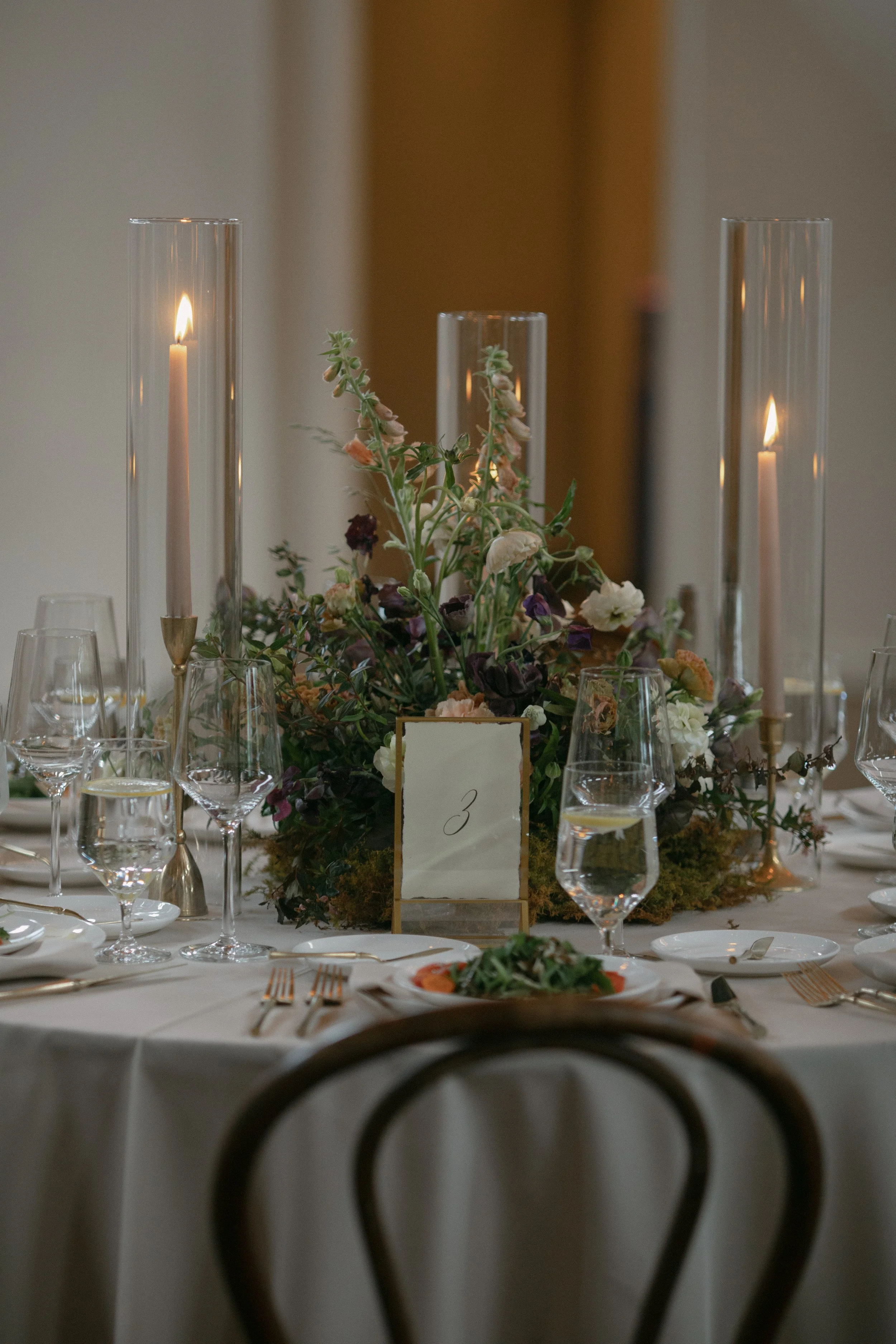 A decorated table at an elegant event, featuring a floral centerpiece, candles in glass holders, wine glasses, plates, and utensils, with a small framed table number sign.