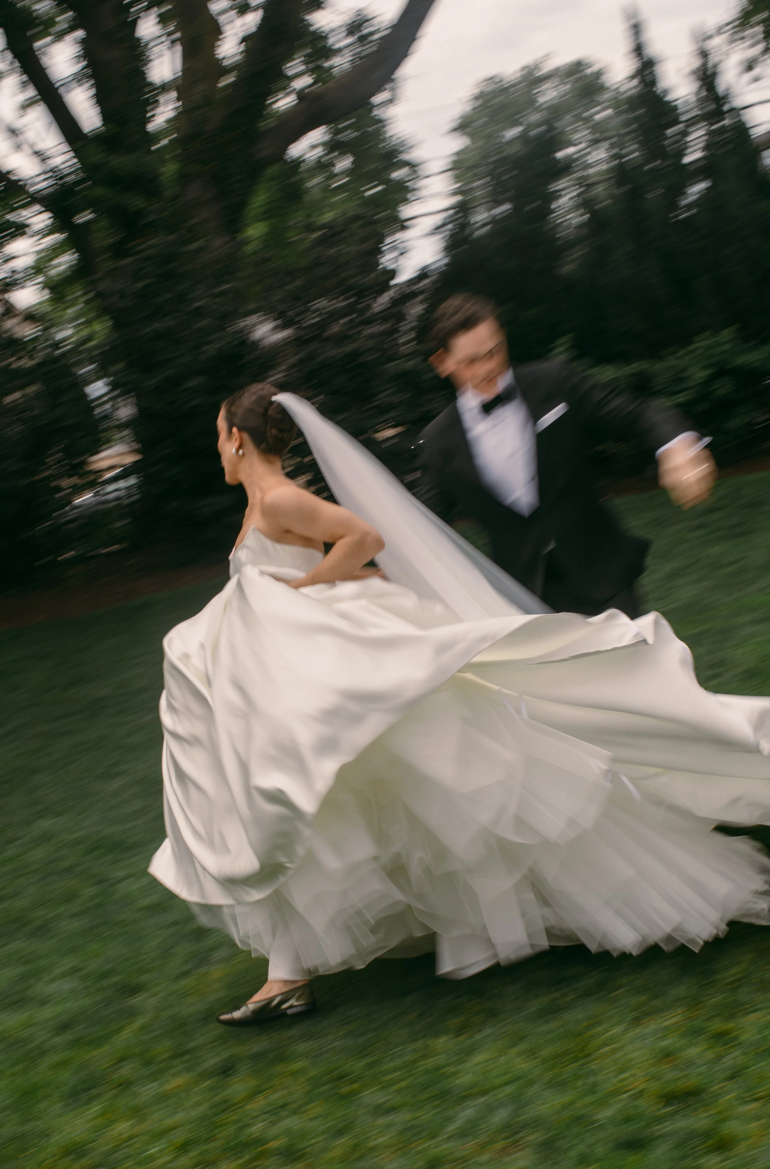 A bride in a white wedding gown running across a grassy area with a groom in a black tuxedo chasing her, blurred background indicating motion.