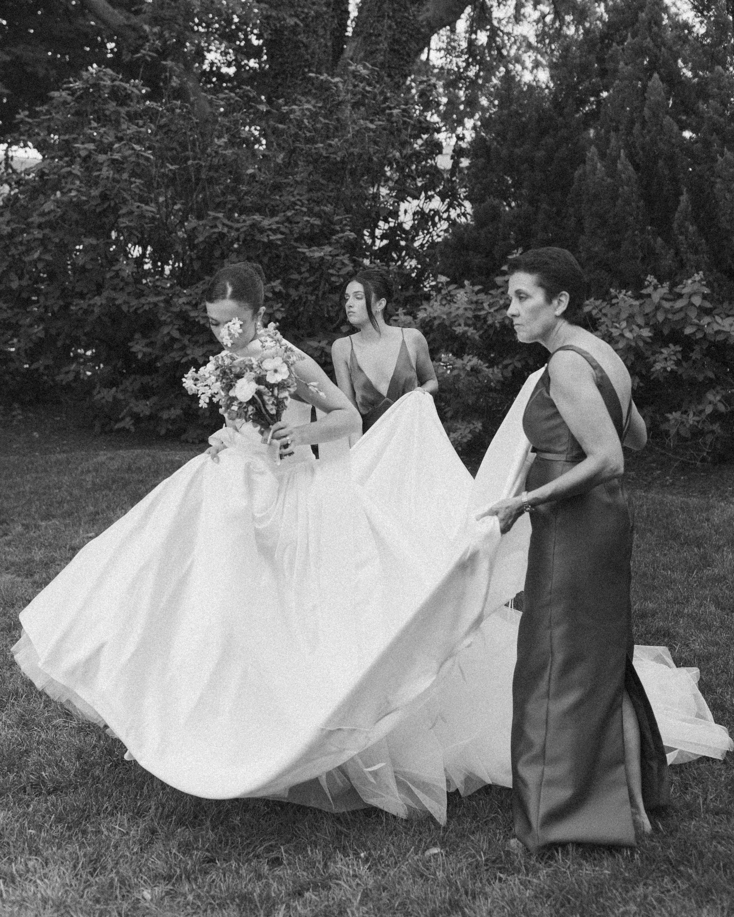 Three women dressed formally outdoors, one holding a bouquet of flowers, with trees in the background.
