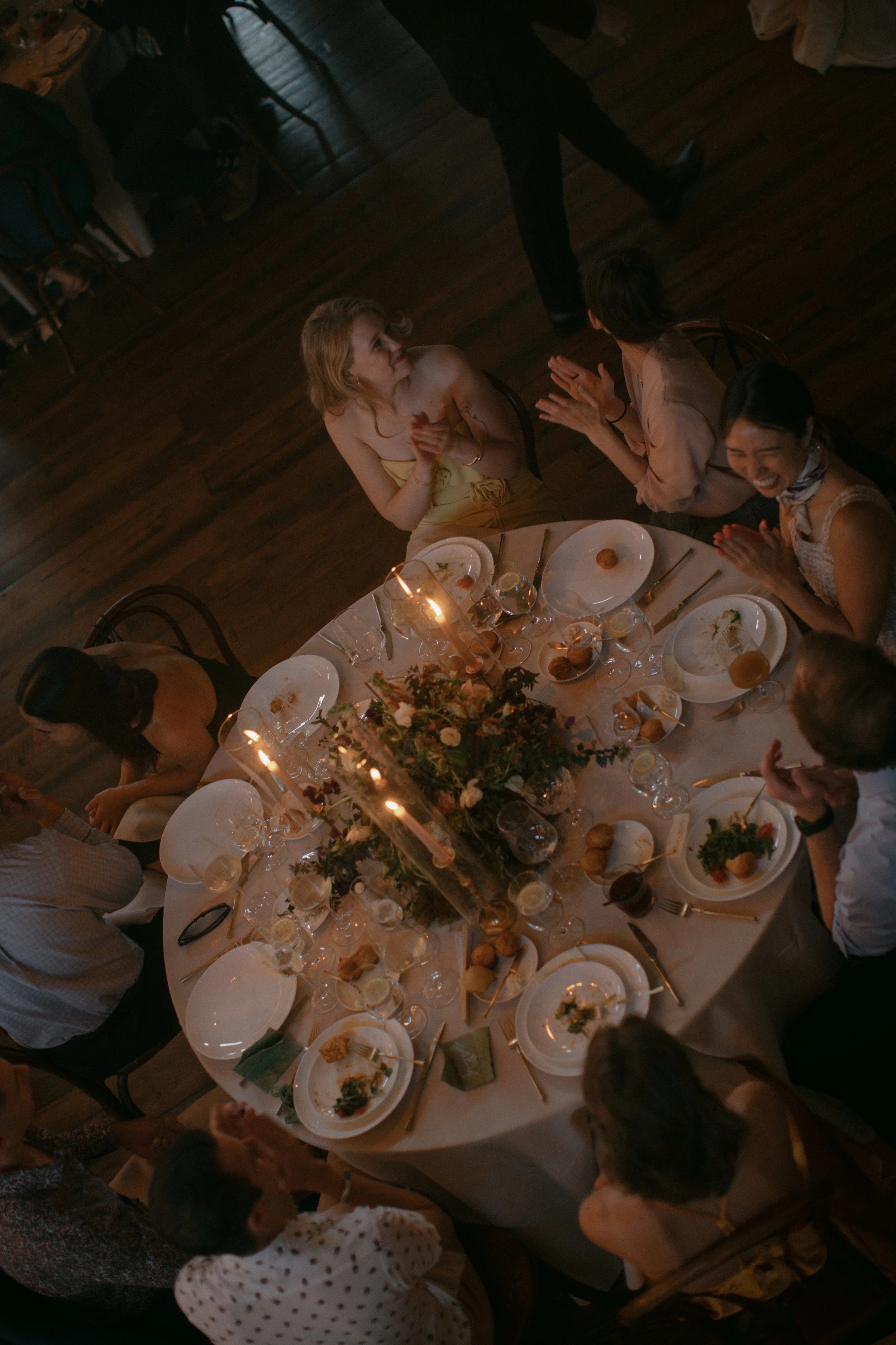 A group of women gathered around a table at a celebration dinner, clapping and smiling. The table is set with plates, glasses, and a floral centerpiece, with some food and drinks visible.