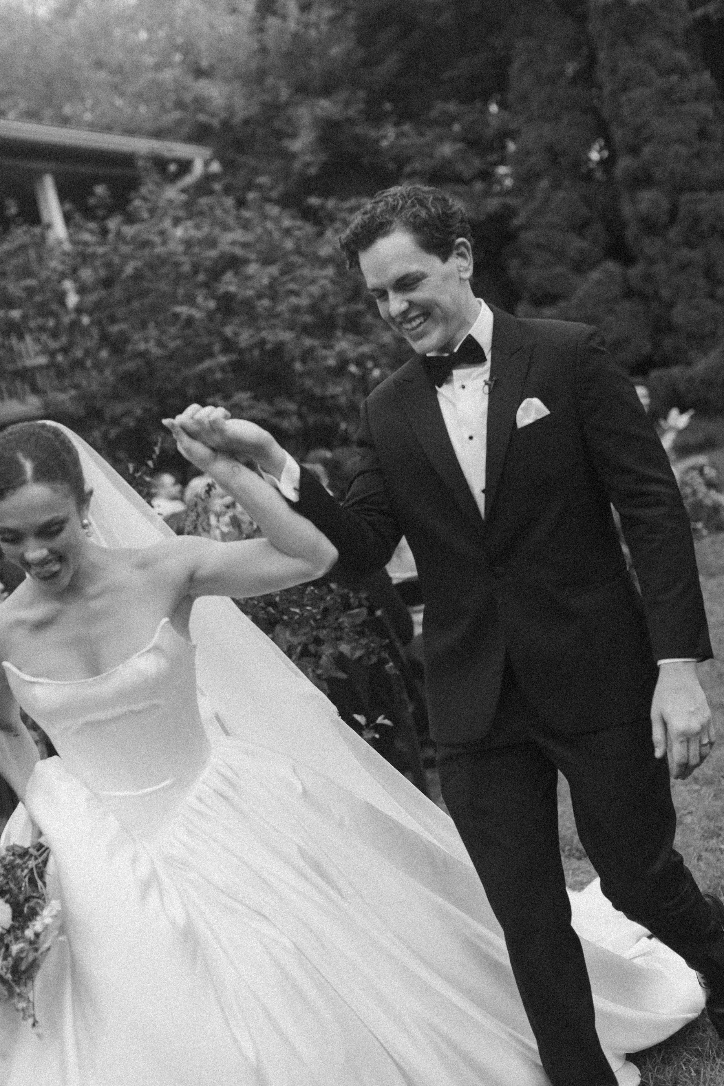 A man in a tuxedo smiling and holding the hand of a woman in a wedding dress, who is standing on a white cloth outdoors with trees and a rock wall in the background.