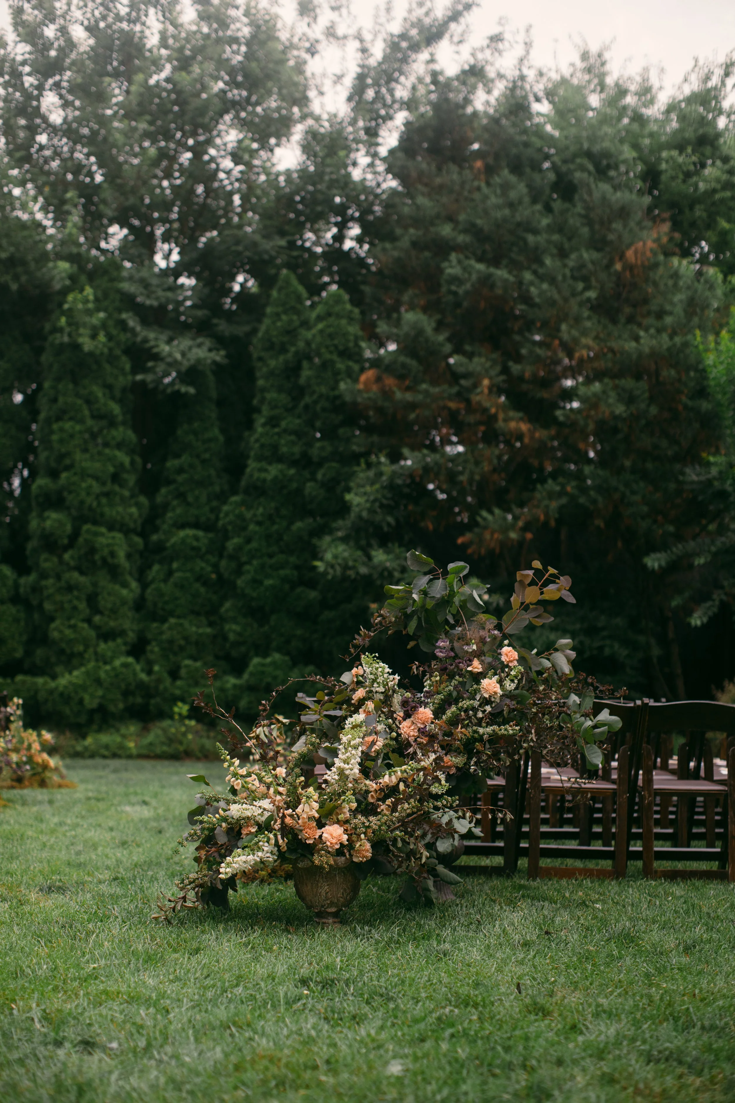 A floral arrangement of pink, white, and purple flowers with leafy greenery in a brown vase on the grass outdoors, with chairs and a large tree in the background.