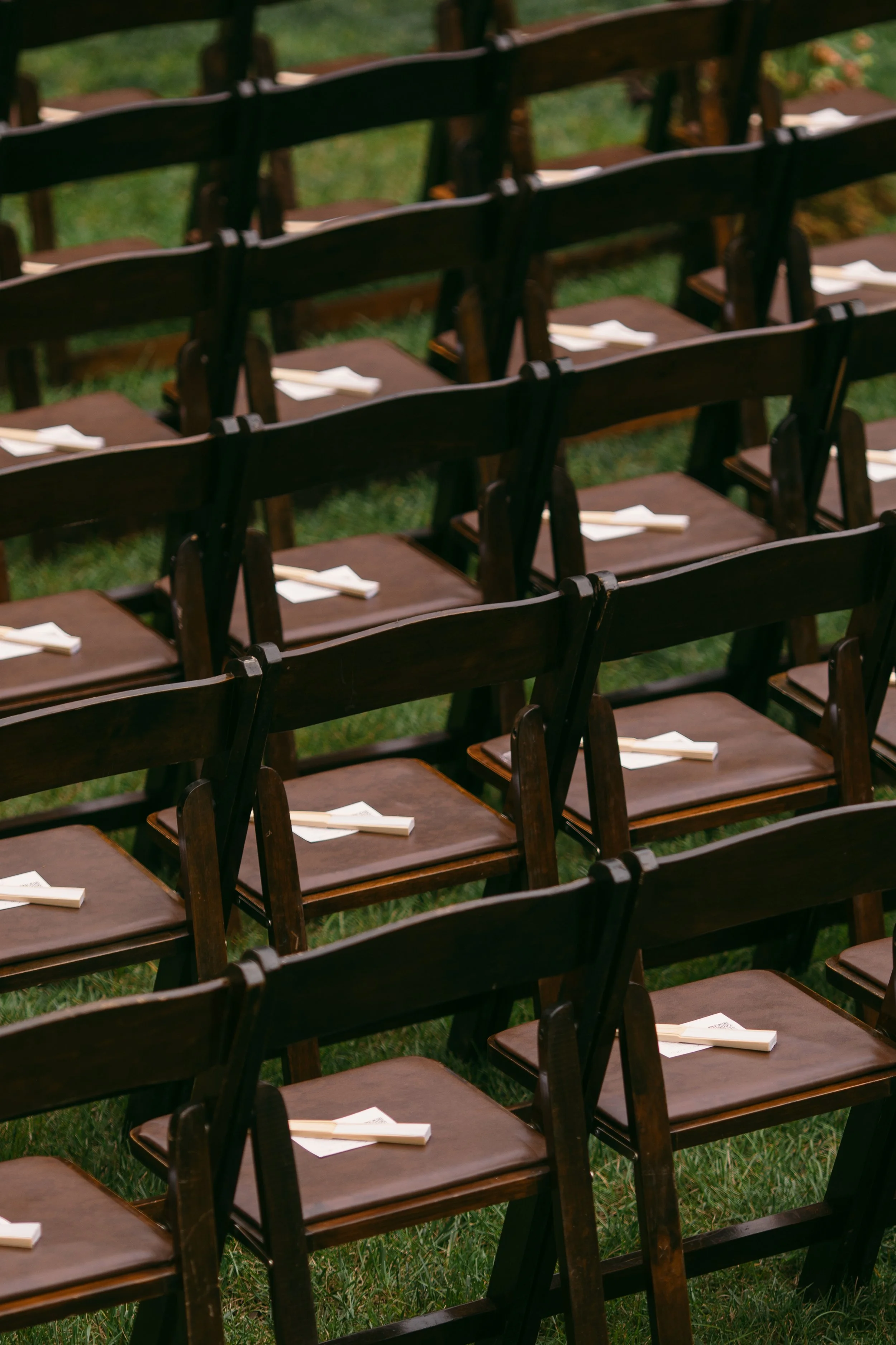 Multiple wooden chairs arranged outdoors on grass, each with a white paper napkin and utensils placed on the seat.