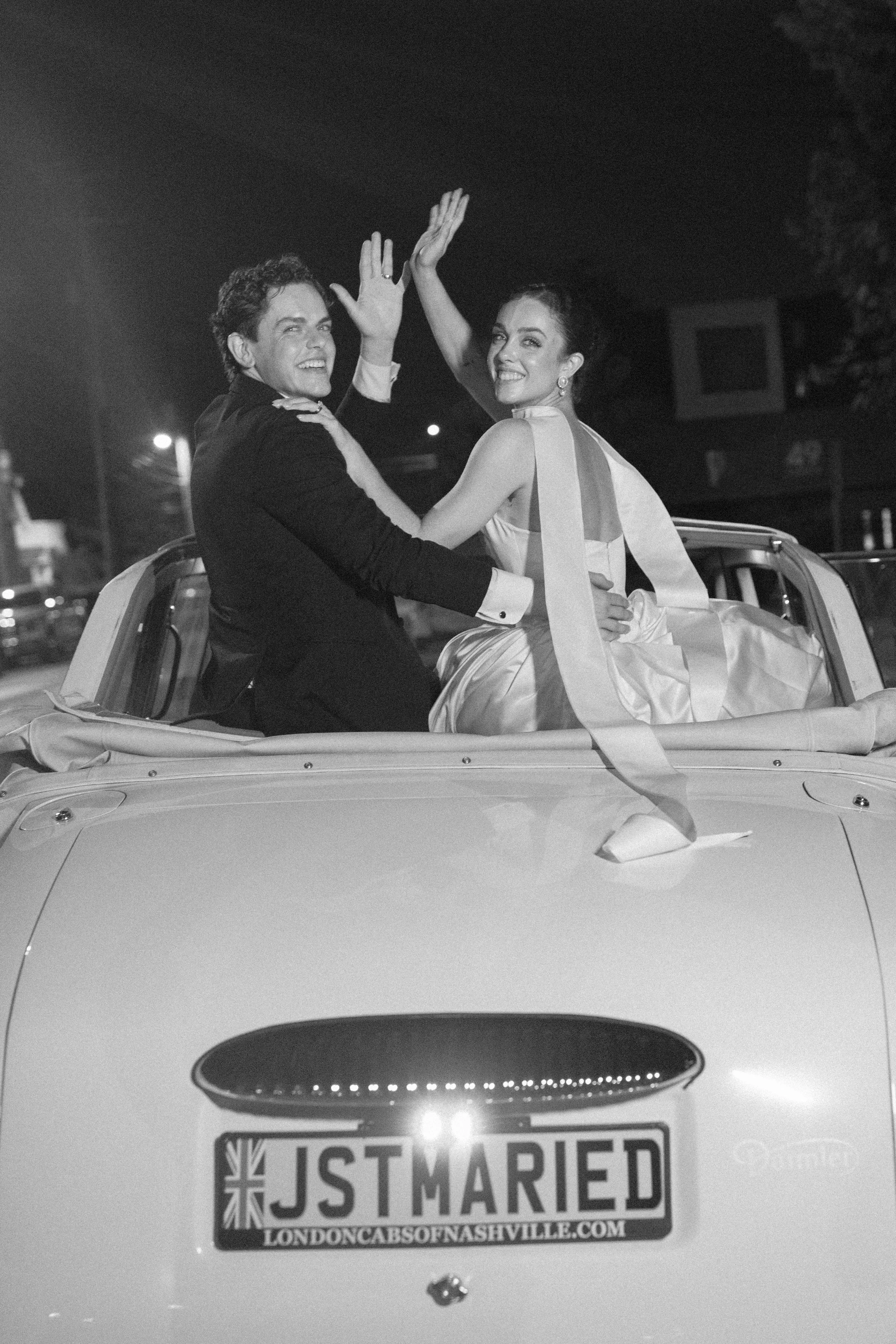 Black and white photo of a newly married couple sitting on the back of a car decorated for their wedding. The man is wearing a suit, and the woman is in a wedding dress with a train. They are smiling and high-fiving each other. The car has a license 