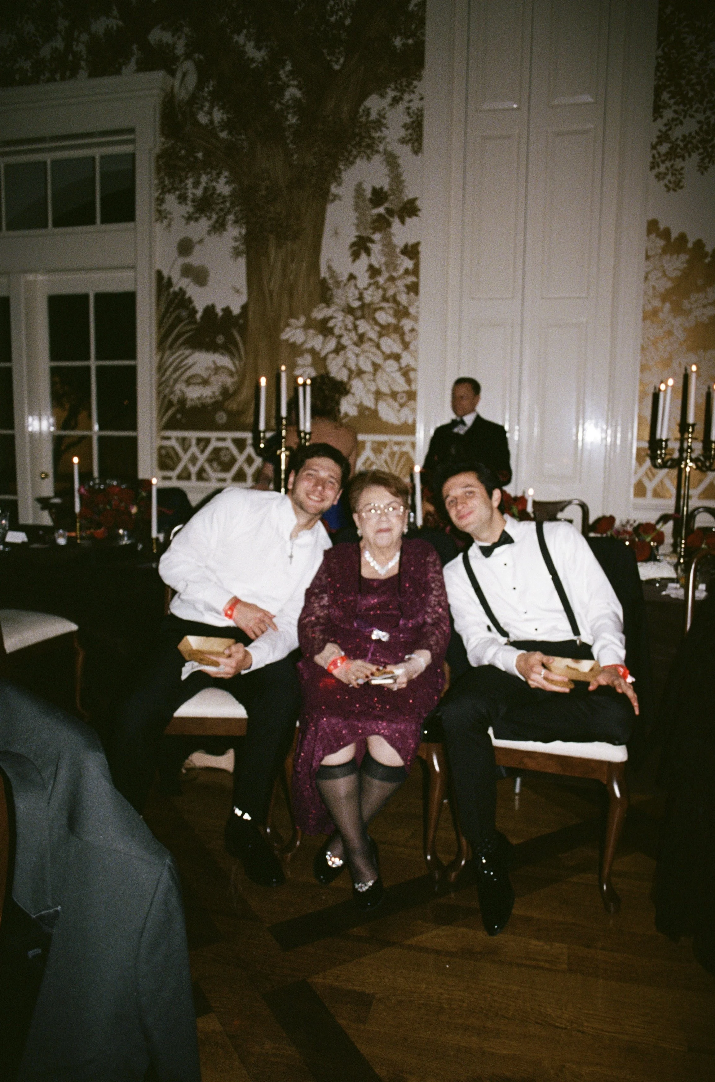 Three people sitting together at a formal event, with two women and one man in tuxedo, and a server standing in the background with a banquet table and floral and candle decorations in the room.
