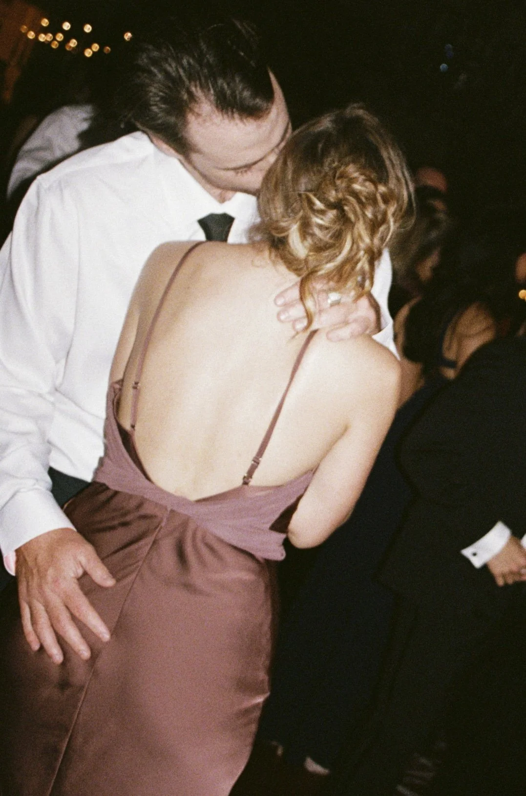 A couple shares a dance kiss at a formal event, the man wearing a white shirt with a black bow tie, and the woman wearing a backless burgundy gown with spaghetti straps, with her hair styled in loose curls.