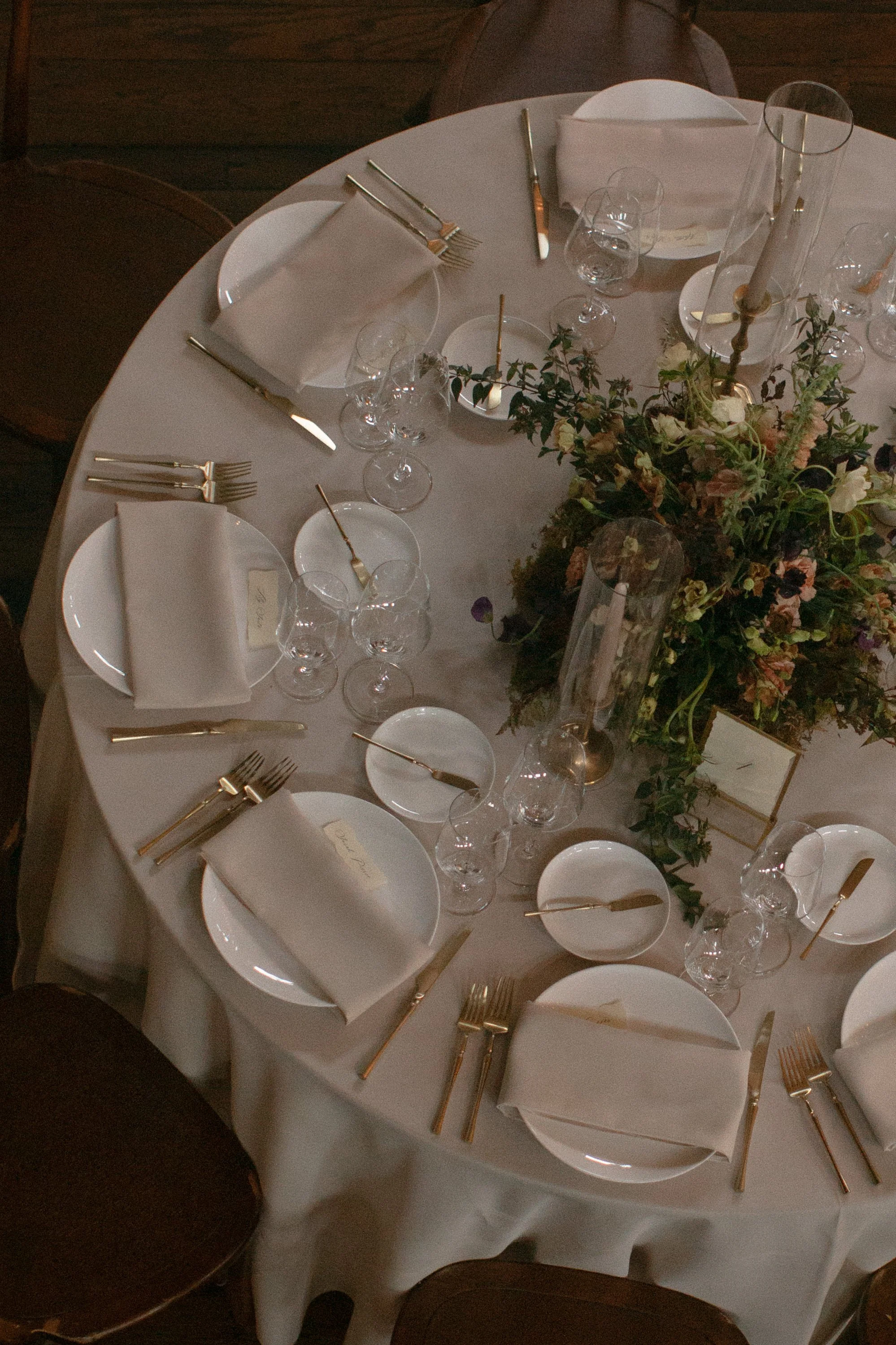 Round dining table set for a formal event with white tablecloth, white plates, gold cutlery, white napkins, clear wine glasses, and a floral centerpiece with greenery and light-colored flowers. The table appears to be in a dimly lit setting.