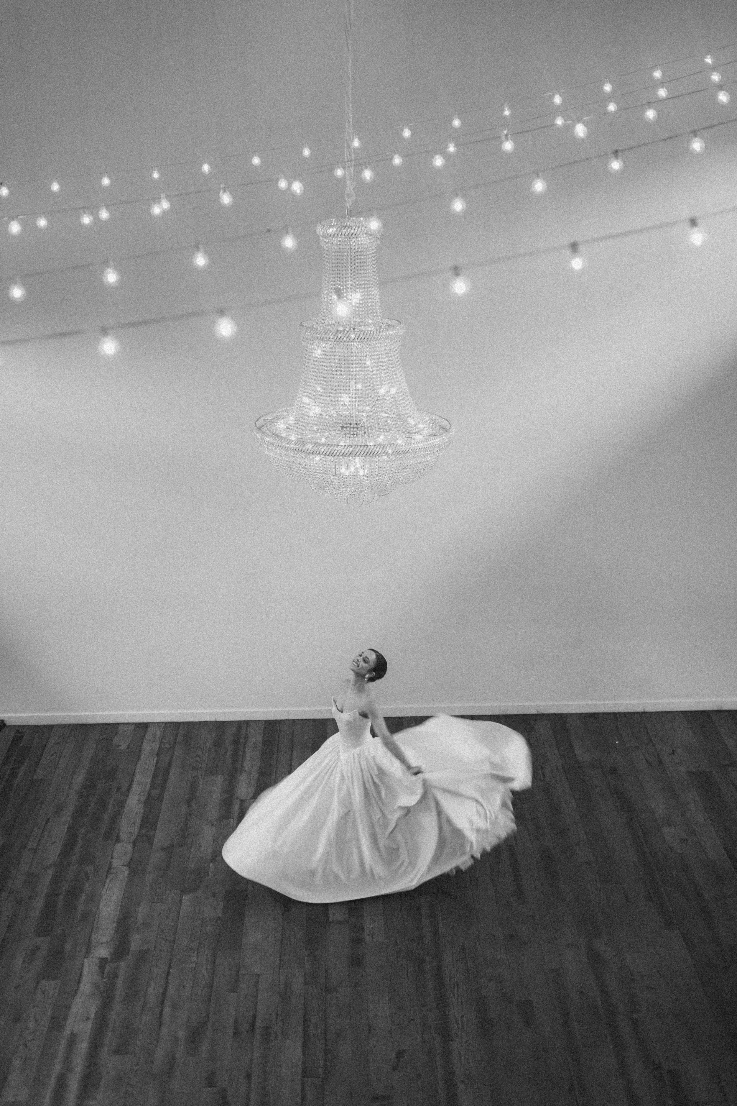 Ballet dancer in a white tutu performing on a wooden floor with a chandelier and string lights above.