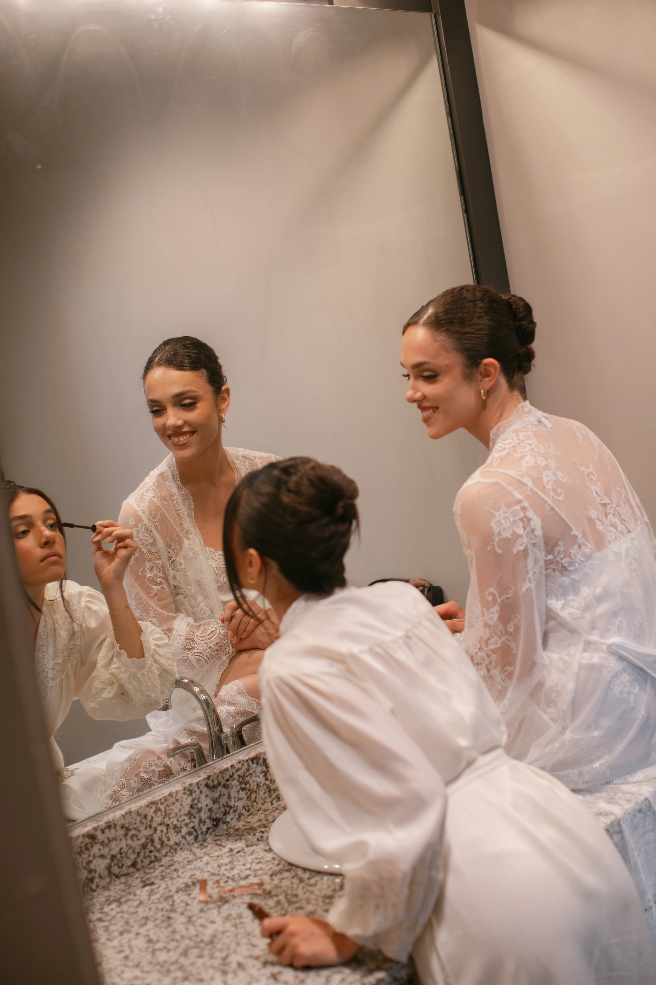 Three women in white lace dresses smiling and applying makeup in front of a mirror in a bathroom with a granite countertop.