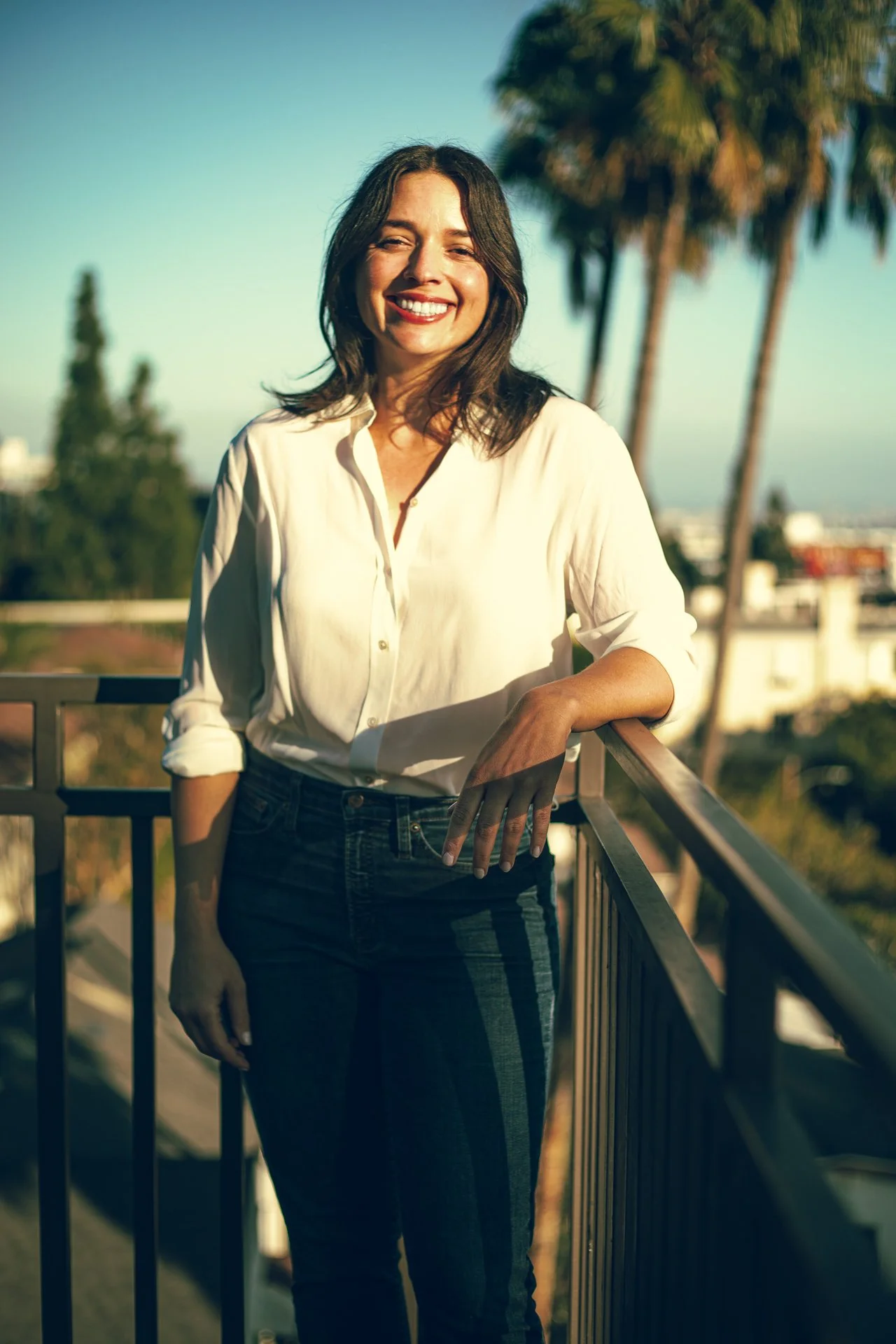 A woman smiling on a balcony during sunset, wearing a white blouse and jeans with shadows cast on her.