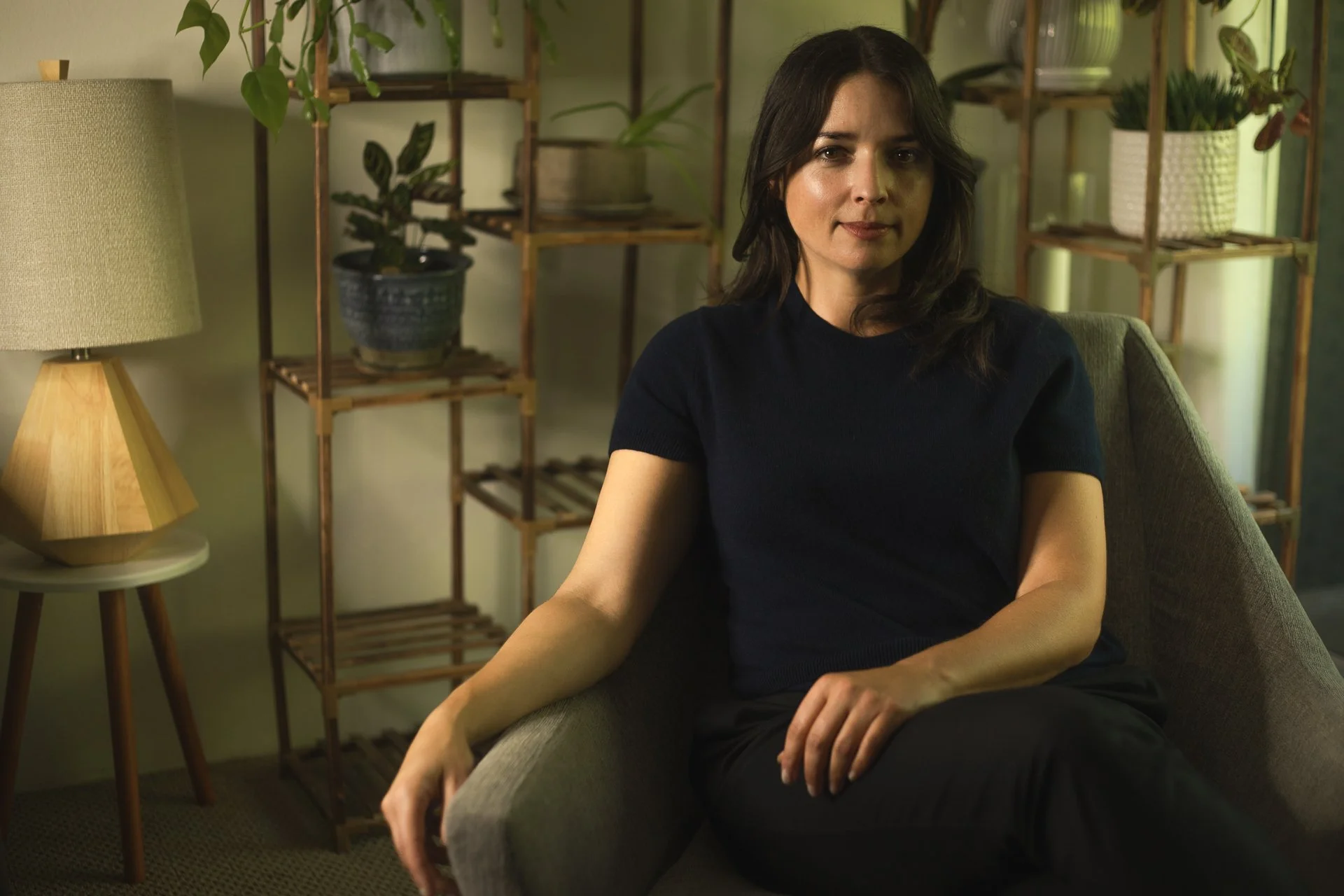 Woman sitting on a gray armchair in a cozy room with indoor plants and wooden shelves in the background, lit softly with a lamp on a side table.