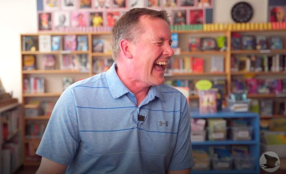 A man laughing and smiling in a room with bookshelves filled with books and comics in the background.