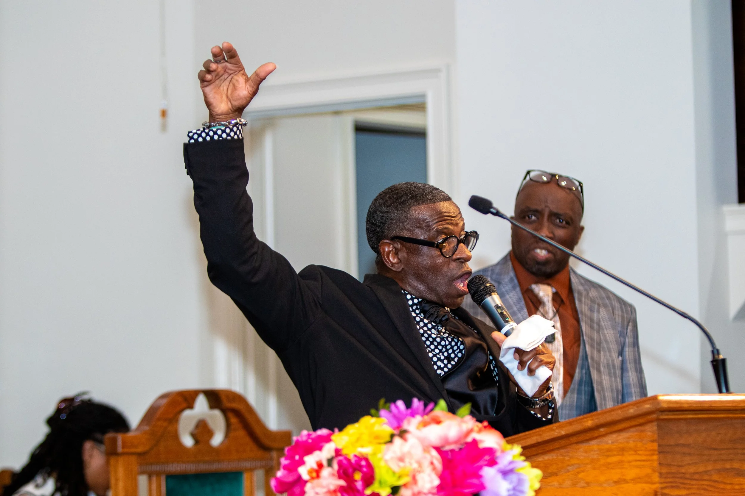 A man with glasses speaking into a microphone, raising his hand, and wearing a black suit with a black and white polka dot shirt, while another man in glasses and a plaid suit listens in the background at a formal event.