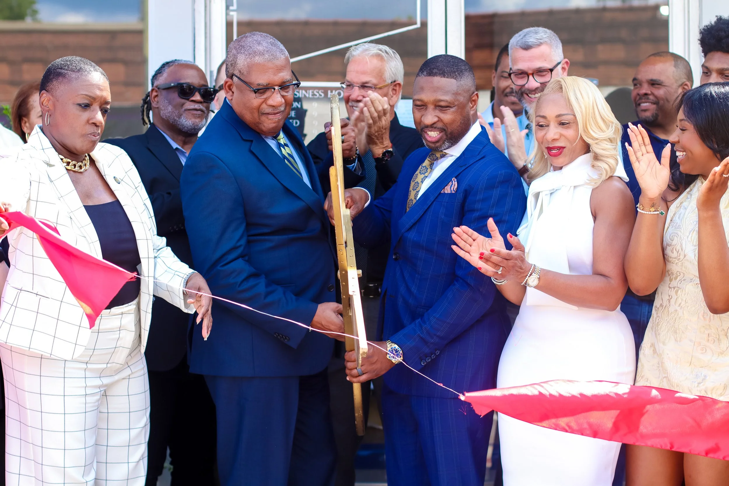 A ribbon-cutting ceremony with a group of diverse adults, some in formal attire, clapping and smiling outside a building.