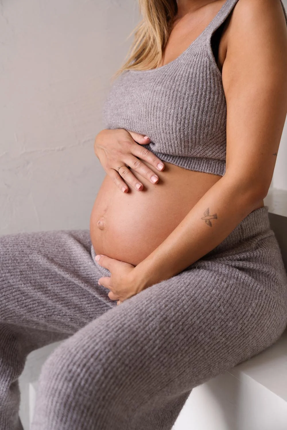 Zwangerschaps shoot in daglichtstudio - Een zwangere vrouw in een grijze top en broek, zittend en haar buik vasthoudend, met een tatoeage op haar onderarm en een ring aan haar vinger.