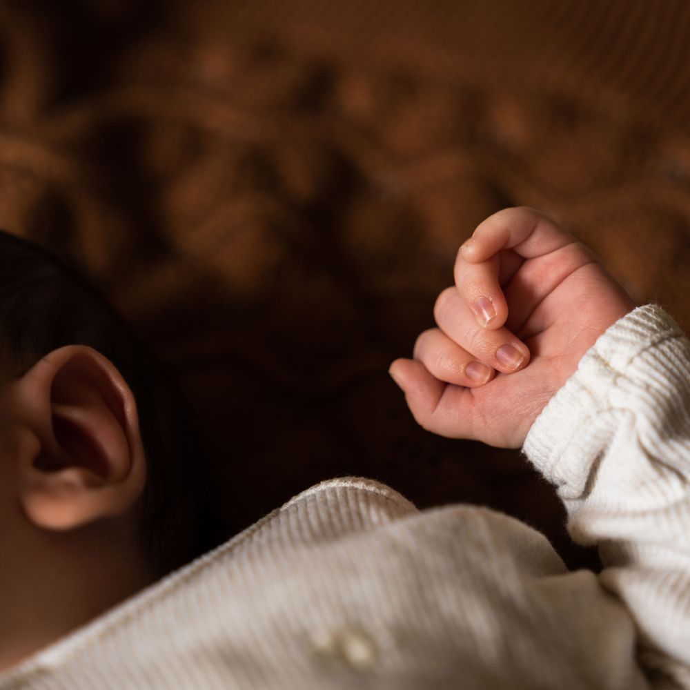 Close-up van een babyhand die een vuistje maakt, met een deel van het hoofd en oor zichtbaar op de linkerzijde, op een zachte, bruine achtergrond. Newborn fotoshoot.