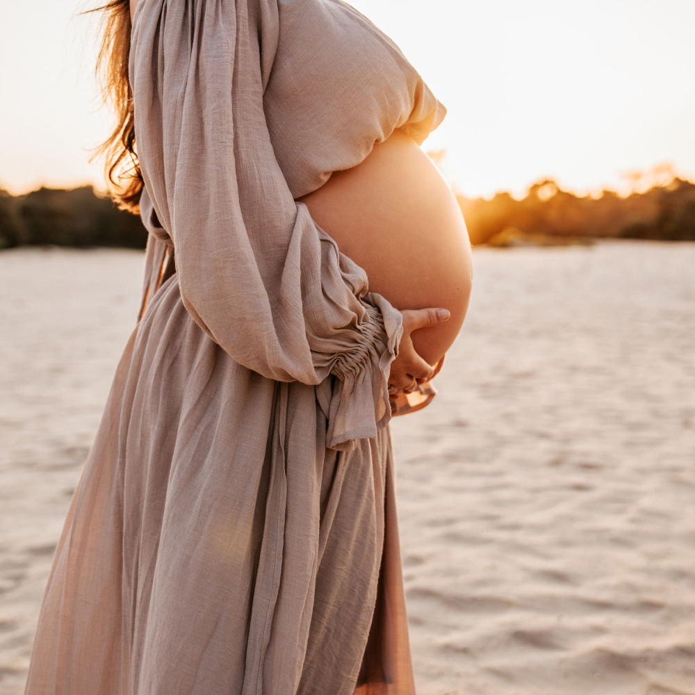 Close-up van een zwangere vrouw die haar buik vasthoudt op het strand tijdens een zwangerschapsshoot bij zonsondergang.