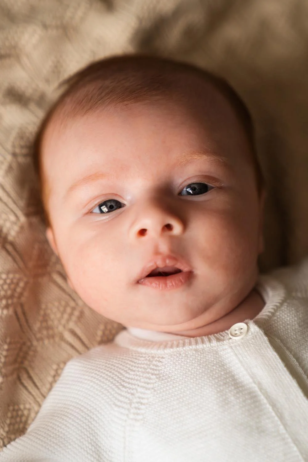 Close-up van een baby met blauwe ogen, liggend op een beige gebreide doek, draagt een witte kledingstuk met knoopje - Newborn shoot.