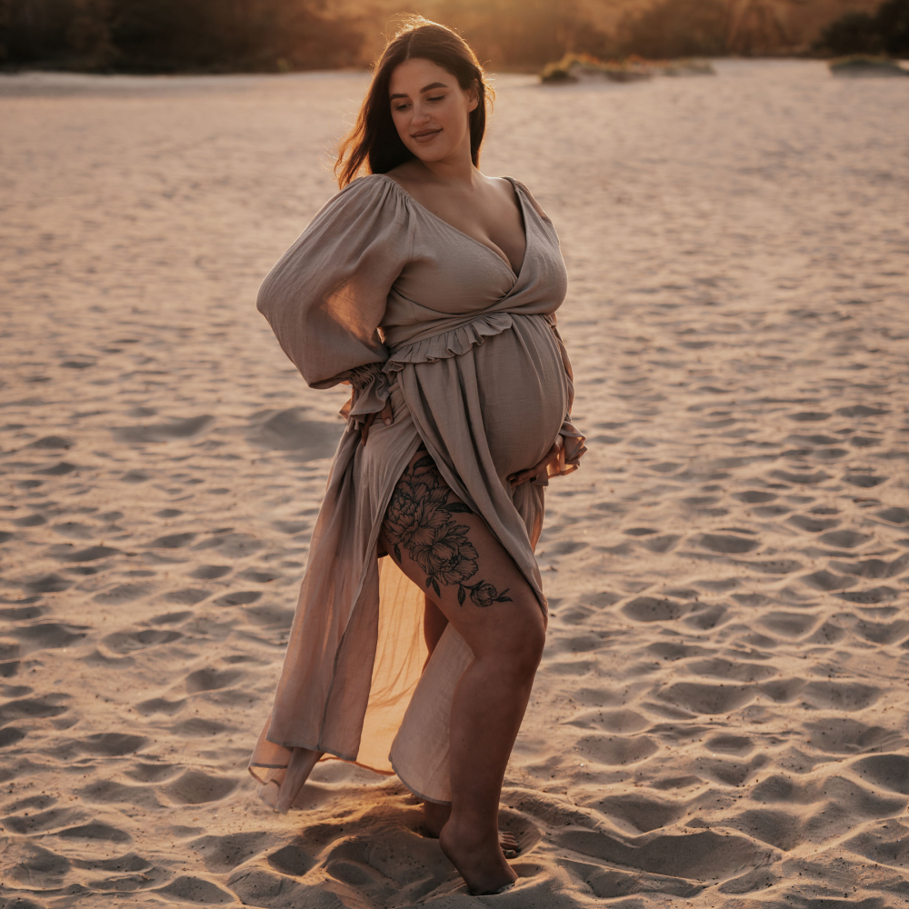 Zwangere vrouw in een beige jurk voor een rivier tijdens de zwangerschapsshoot met zonsondergang.