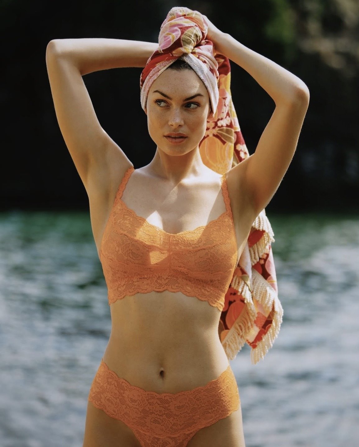 A woman standing outdoors near water, wearing an orange lace bralette and matching underwear, with a colorful towel wrapped around her head.