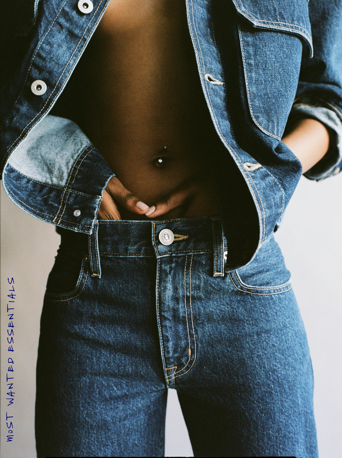 Close-up of a person wearing a denim jacket and jeans, with their shirt lifted to expose their midriff and belly button piercing.