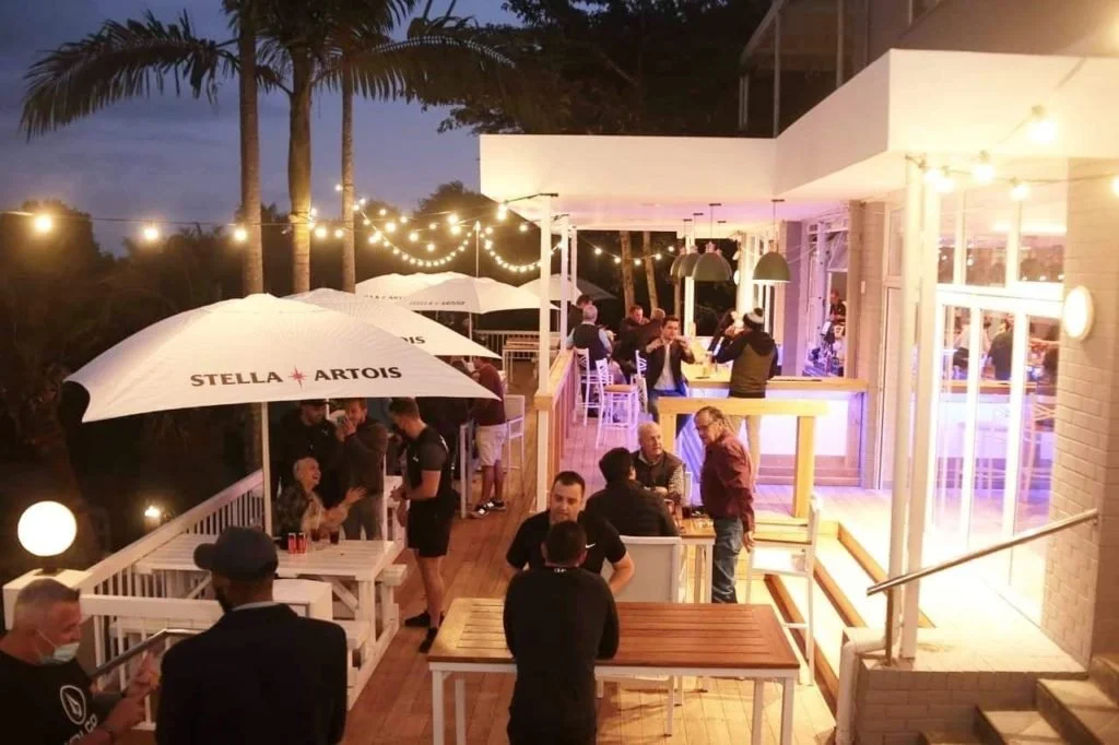 Buitenverhuur met groepen mensen bij een restaurant of bar, met parasols, festoon verlichting, en palmbomen, in de avond.