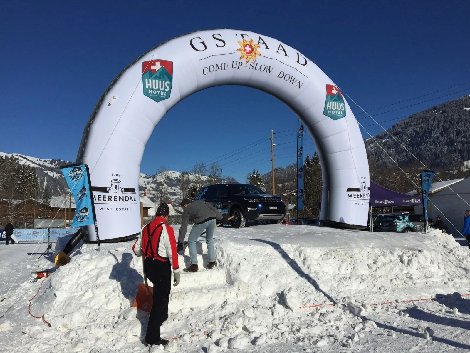 Auto op een kleine verhoogde sneeuwbank onder een grote witte boog met teksten en logo's, personen die de sneeuwbank aanpassen, in een sneeuwrijke omgeving met bergen en een blauwe lucht.