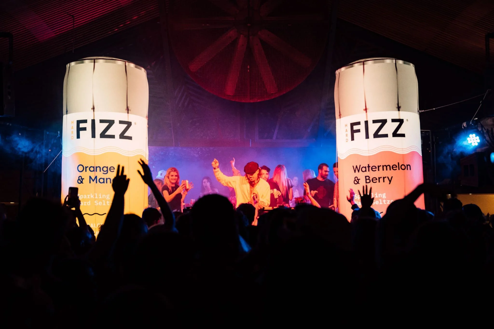 Een druk muziekfestival met een dansfeest in een grote zaal, mensen in silhouet voor het podium, met verlichte borden die FIZZ en smaken zoals Orange & Mango en Watermelon & Berry tonen.