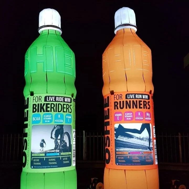 Opblaasbare advertenties voor sportdrankjes, een voor bikers en een voor runners, 'live ride win' en 'live run win' in een donker kader in de nacht.