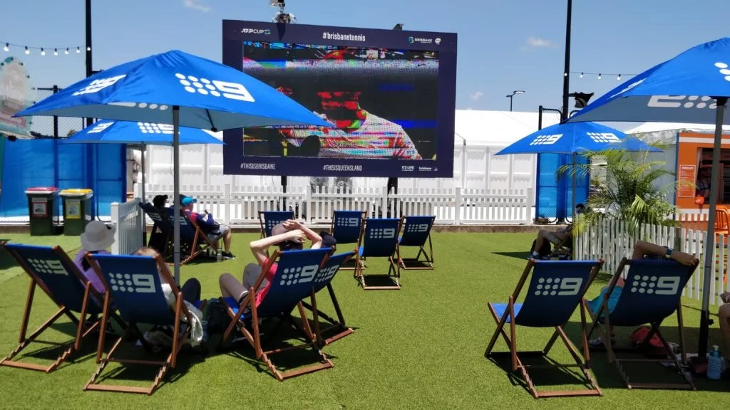 Mensen kijken naar een groot buitenscherm op een grasveld met parasols, zitplaatsen en een witte schutmuur