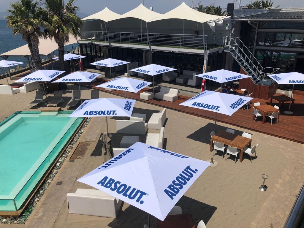 Zonnige strandlocatie met ligbedden en tafels onder witte parasols met het logo van Absolut. Er is een klein zwembad en een terras met uitzicht op het water.