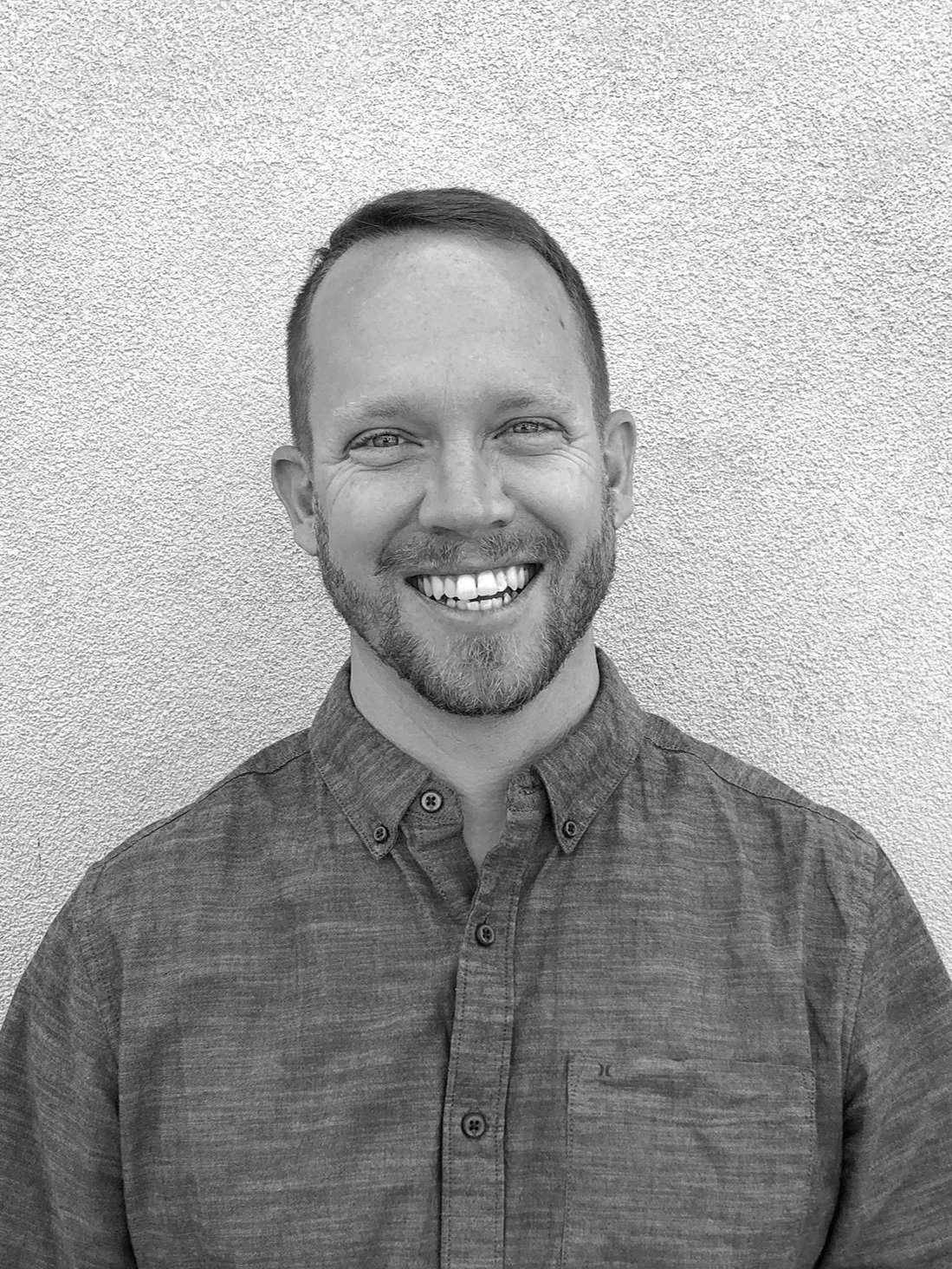 A smiling man with a beard and short hair, wearing a button-up shirt, standing against a textured wall.