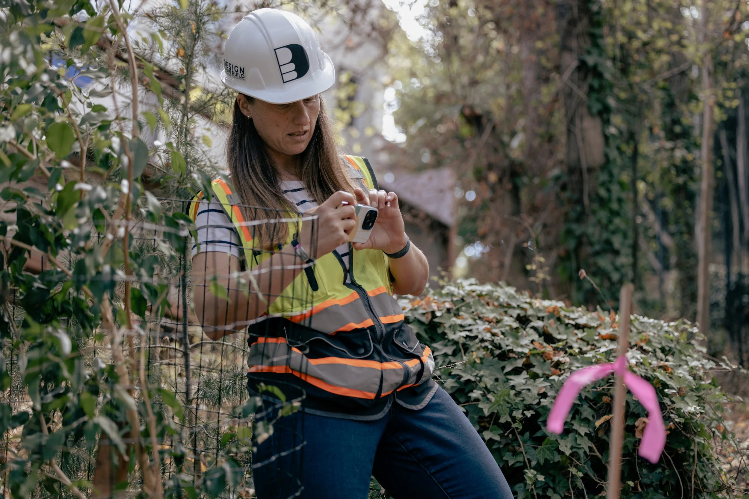 A woman wearing a safety helmet and a reflective safety vest performing site surveys for client property lines. 