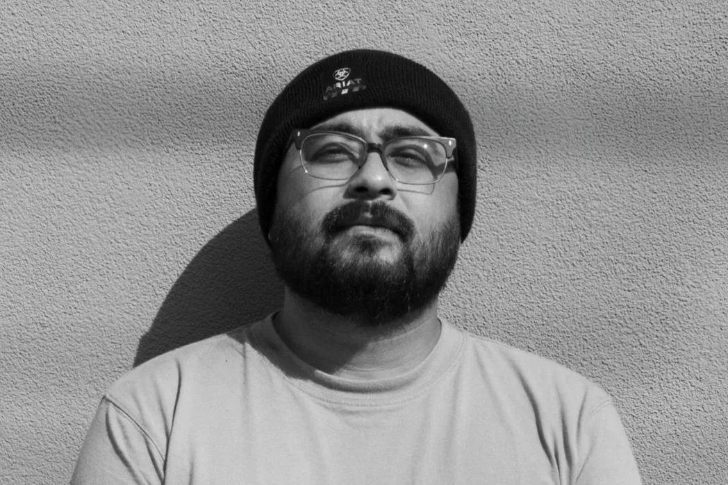 Black and white photo of a man with glasses and a beard, wearing a beanie, standing against a textured wall.