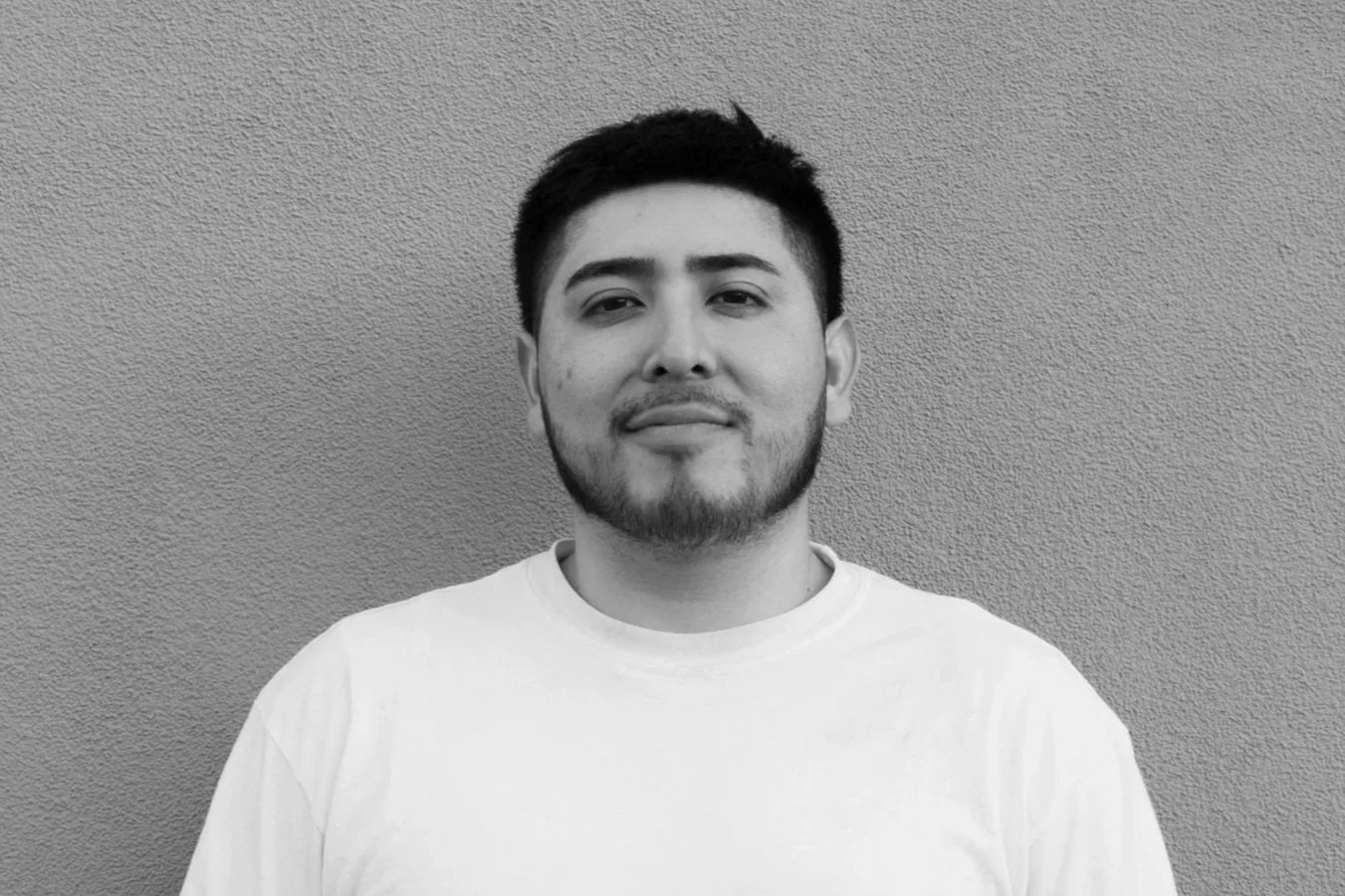Black and white photo of a young man with short dark hair, light facial hair, wearing a white shirt, standing against a textured wall.