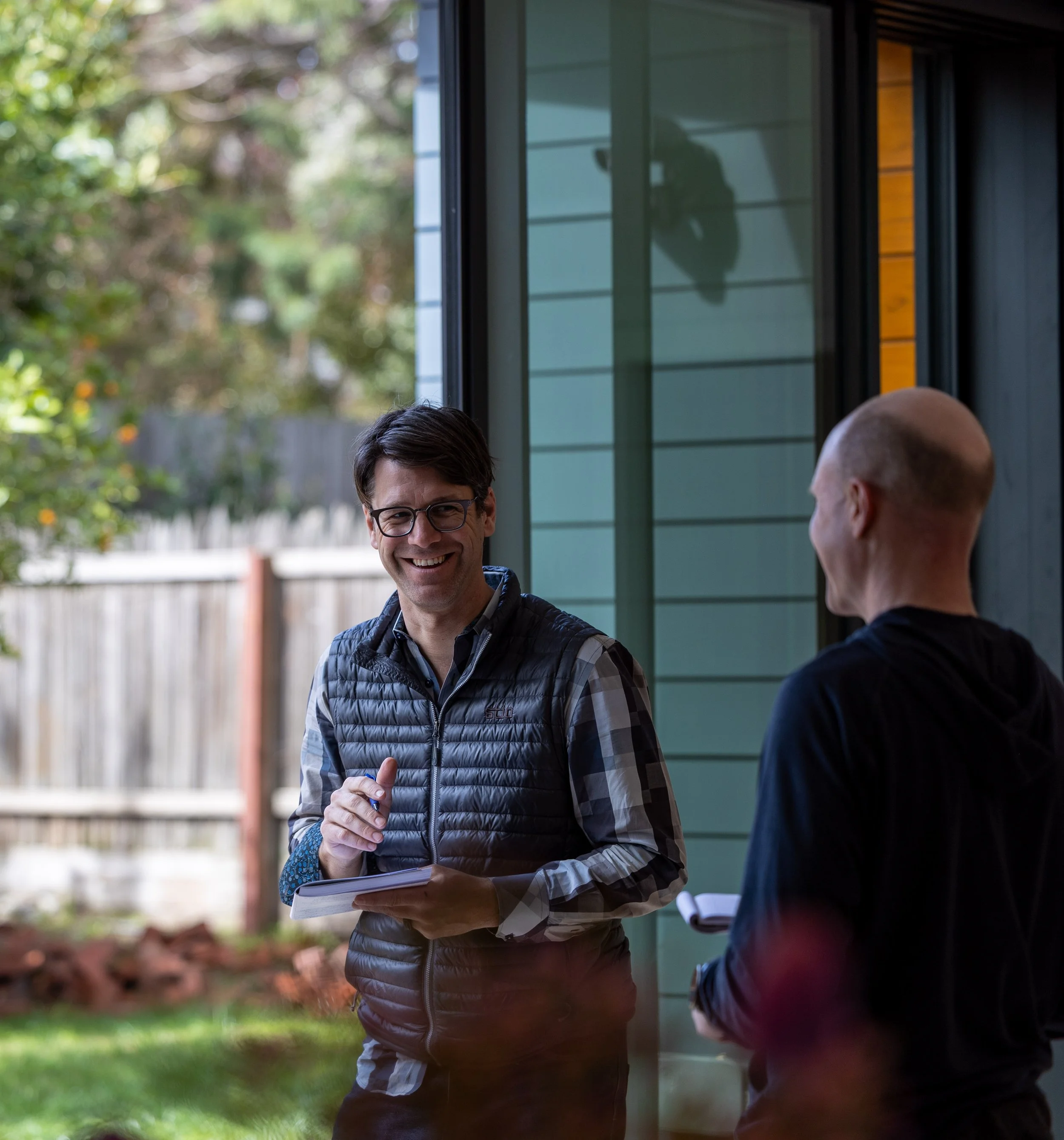 Two men having a conversation outdoors near a modern house. One is smiling and holding a notebook, the other is partially visible and facing away. Trees and a fence are in the background.