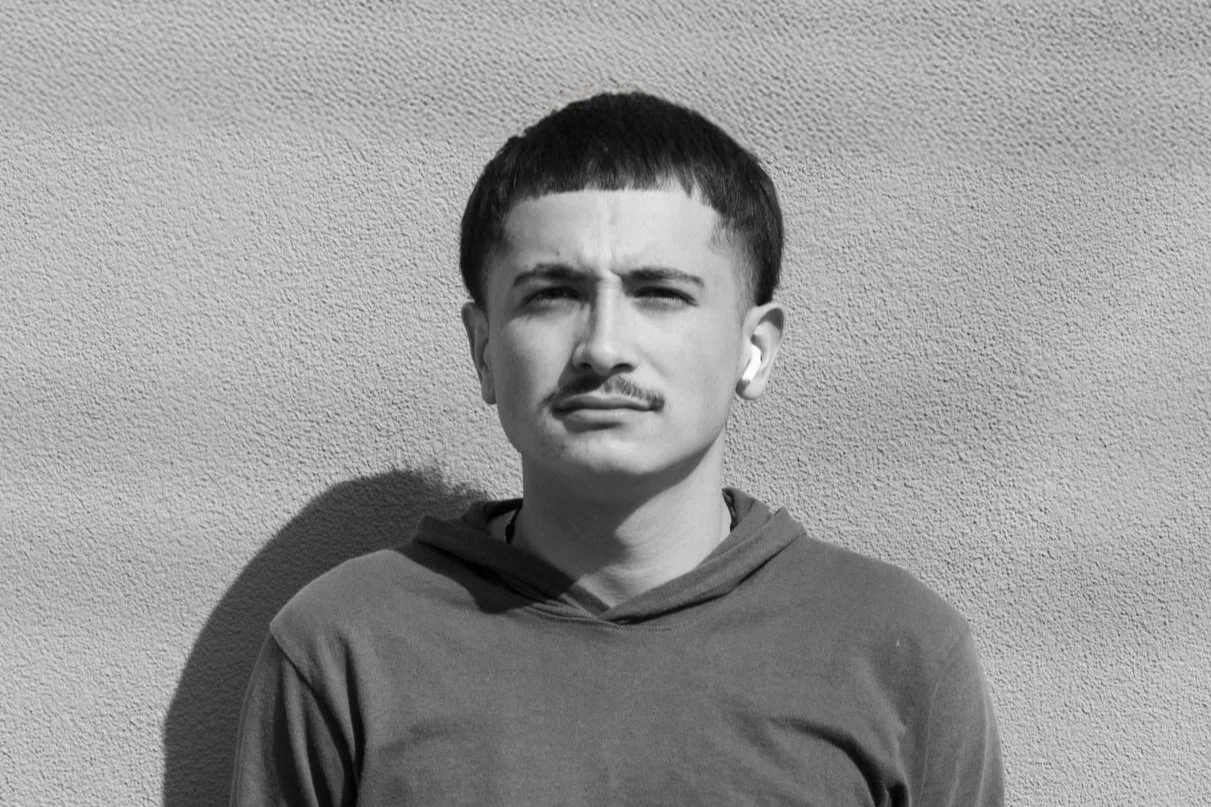 A young man with short dark hair and a mustache, wearing a hoodie and wireless earbuds, standing against a textured wall in sunlight.
