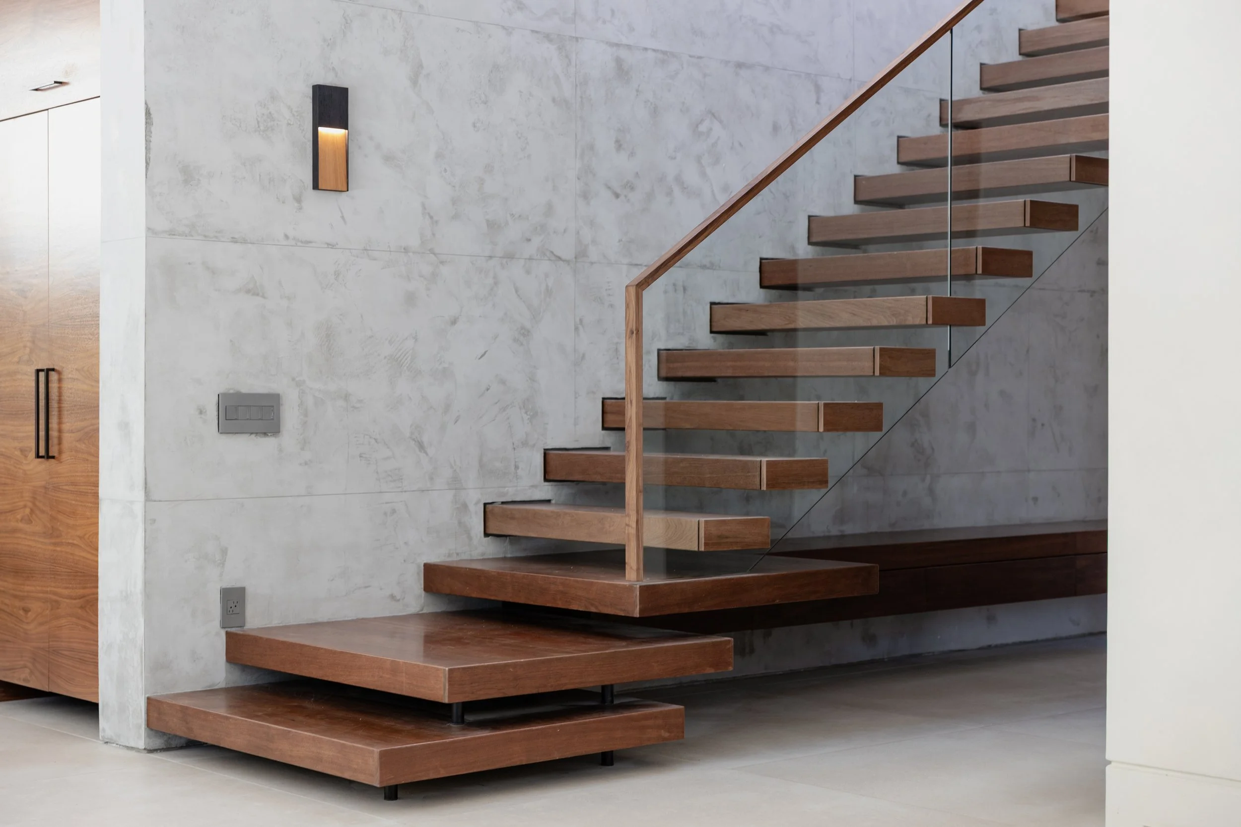 A concrete wall of a modern staircase with wooden steps, illuminated by skylights, casting a rainbow reflection on the wall.