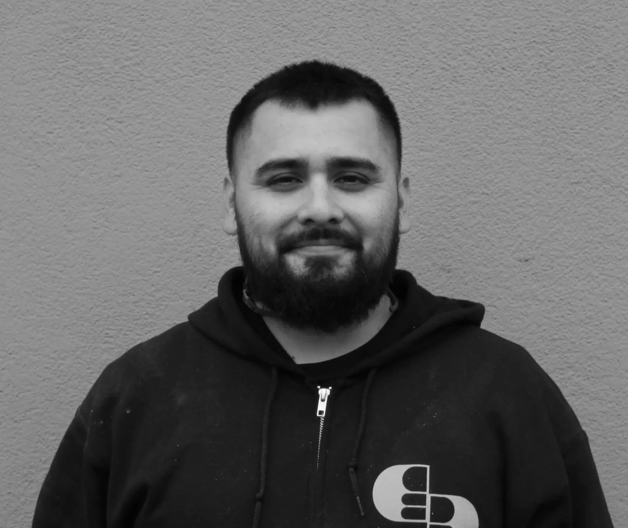 Black and white photo of a young man with short hair and a beard, wearing a dark hoodie with a zipper, standing against a textured wall.