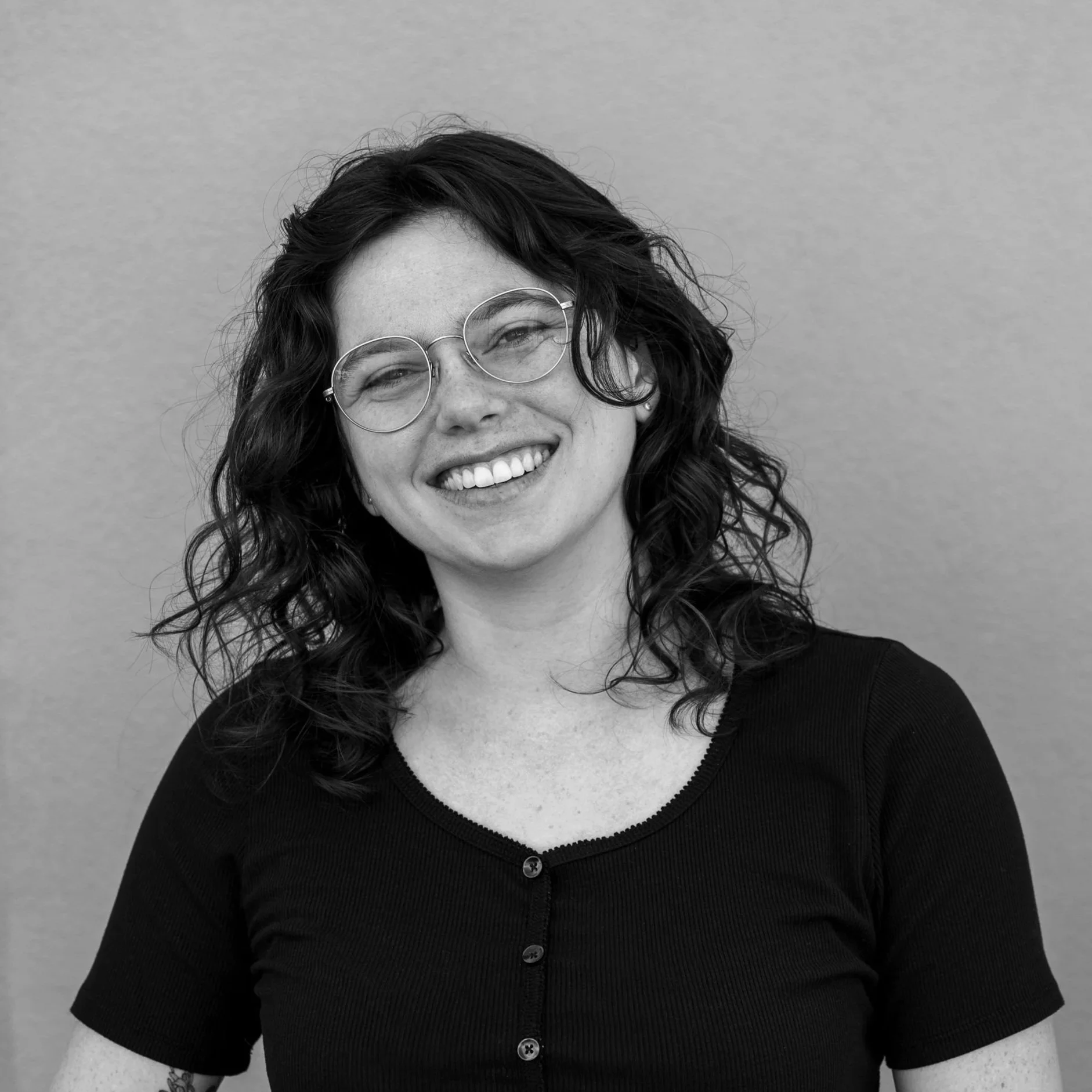 Close-up black-and-white portrait of a woman with curly hair, wearing glasses, smiling, and dressed in a black button-up shirt.