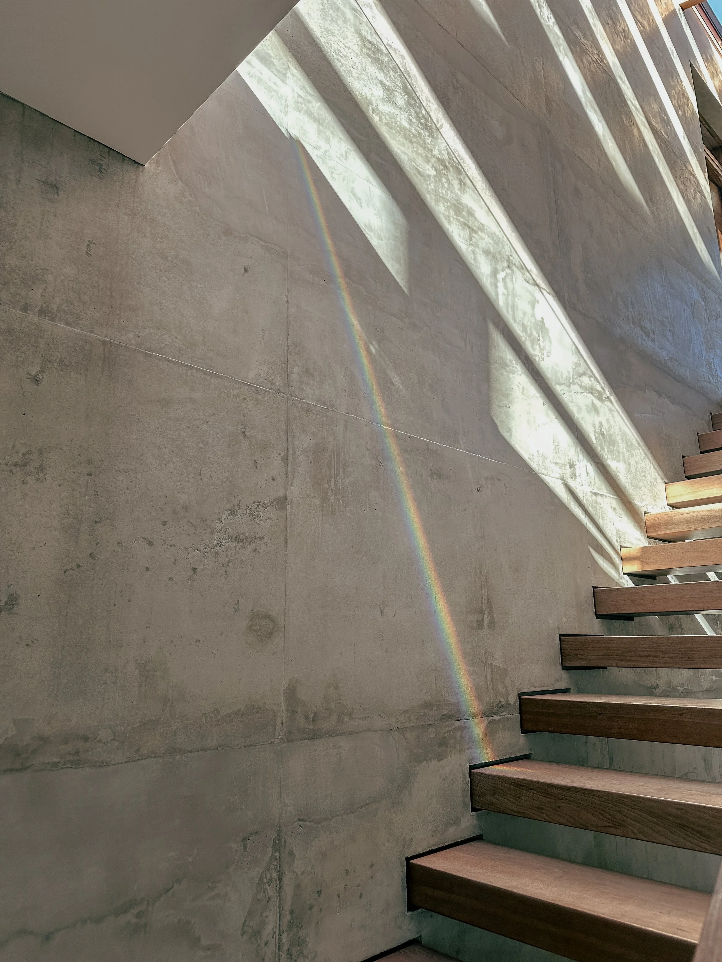 A concrete wall of a modern staircase with wooden steps, illuminated by sunlight, casting a rainbow reflection on the wall.