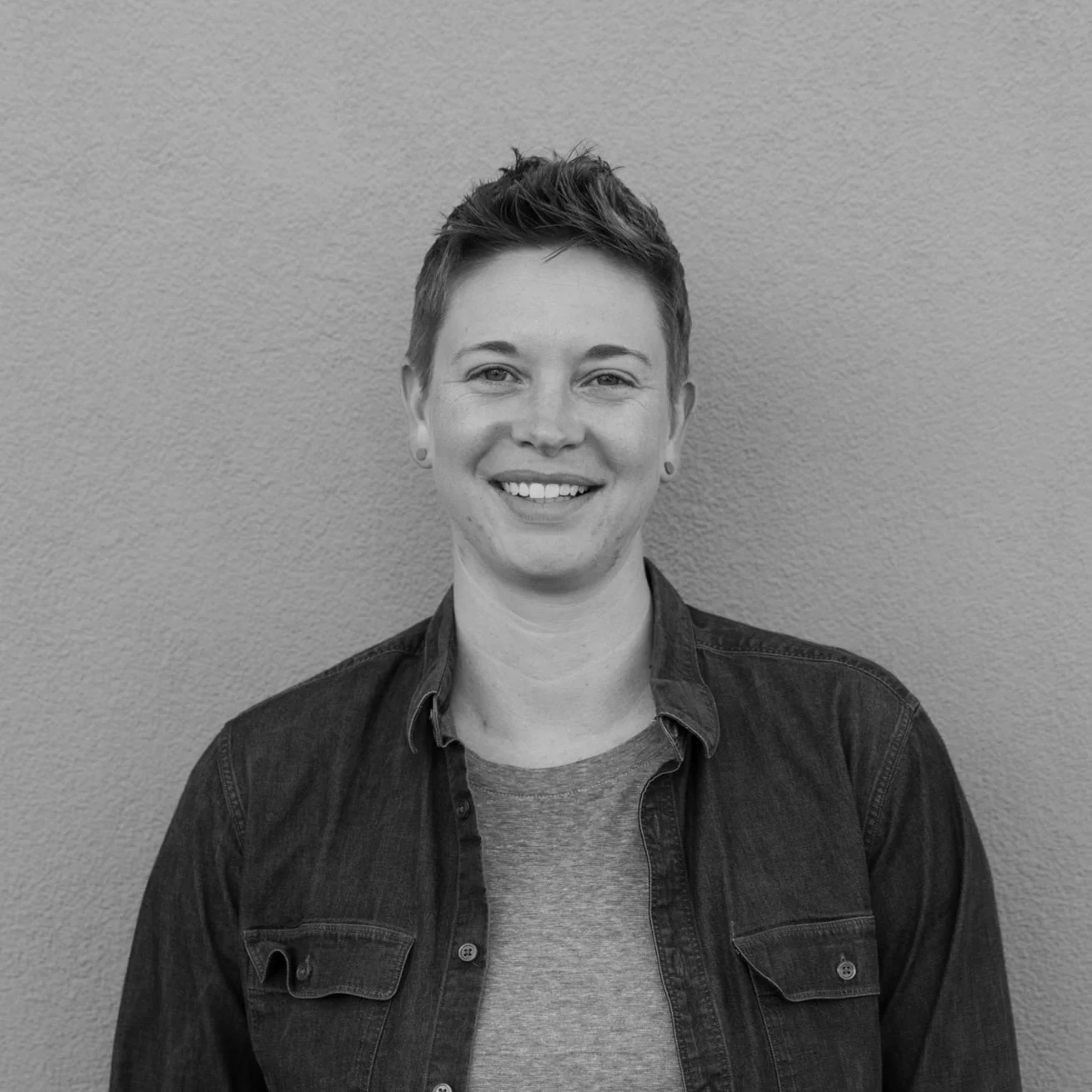 Black and white portrait of a smiling woman with short hair, wearing a denim shirt over a T-shirt, standing against a plain wall.